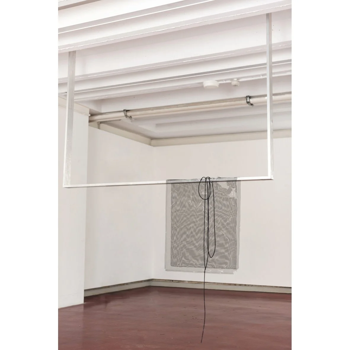 Art installation featuring a large geometric metal frame hanging in an empty white gallery space with polished wooden floors, a mesh screen, and exposed ceiling pipes.