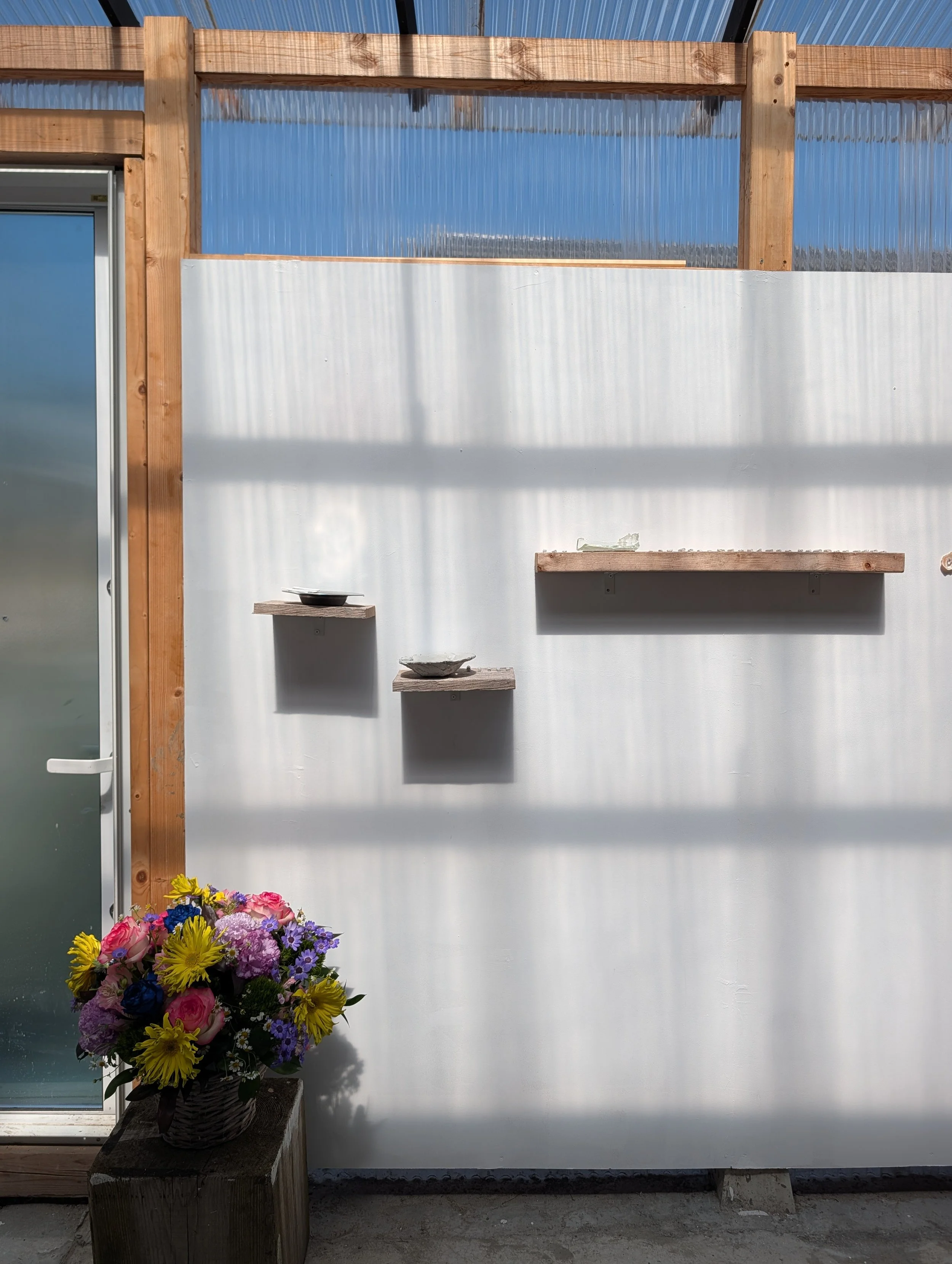 A large colorful flower arrangement with pink, purple, blue, and yellow flowers placed on a wooden bench near a white wall. There are three floating shelves on the white wall above the flower arrangement and colorful wooden beams framing part of the 