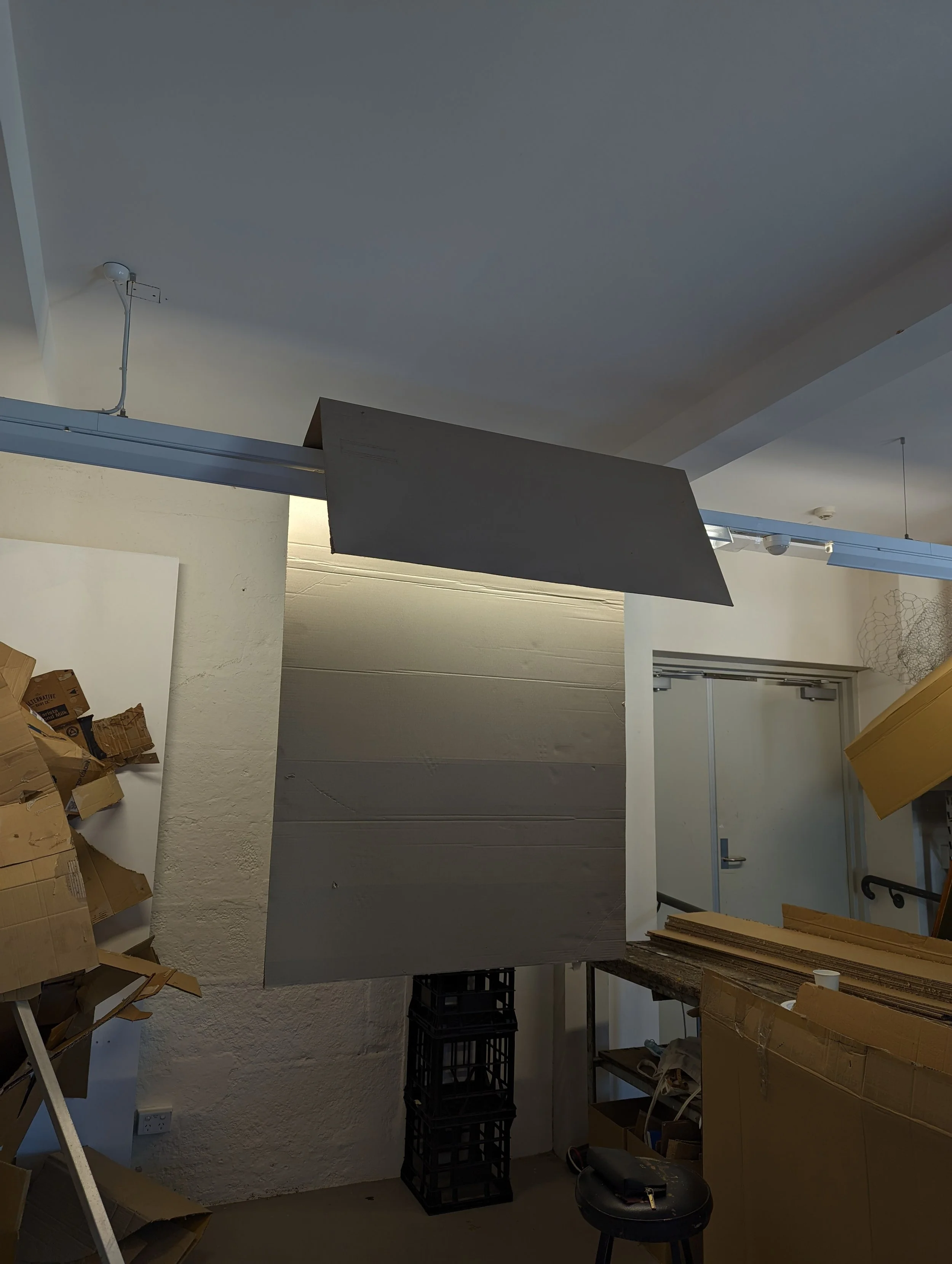 A storage or workspace area with cardboard boxes, a black plastic crate, a small black table, and a cabinet with boxes on top. The ceiling has an exposed light fixture, and the door in the background is closed.