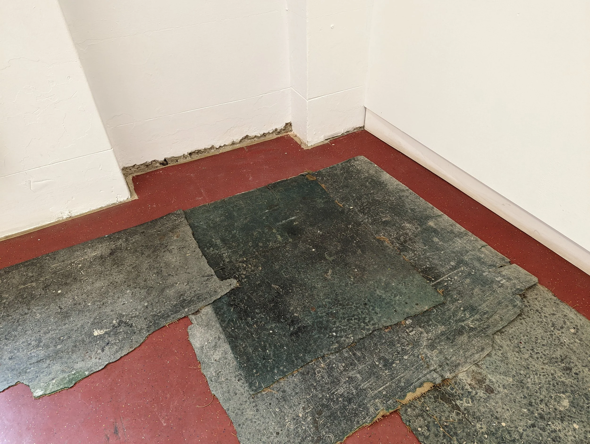 Close-up of a corner of a room showing peeling paint on white walls, a red and black worn-out floor, and a piece of black rubber or foam mat on the floor.
