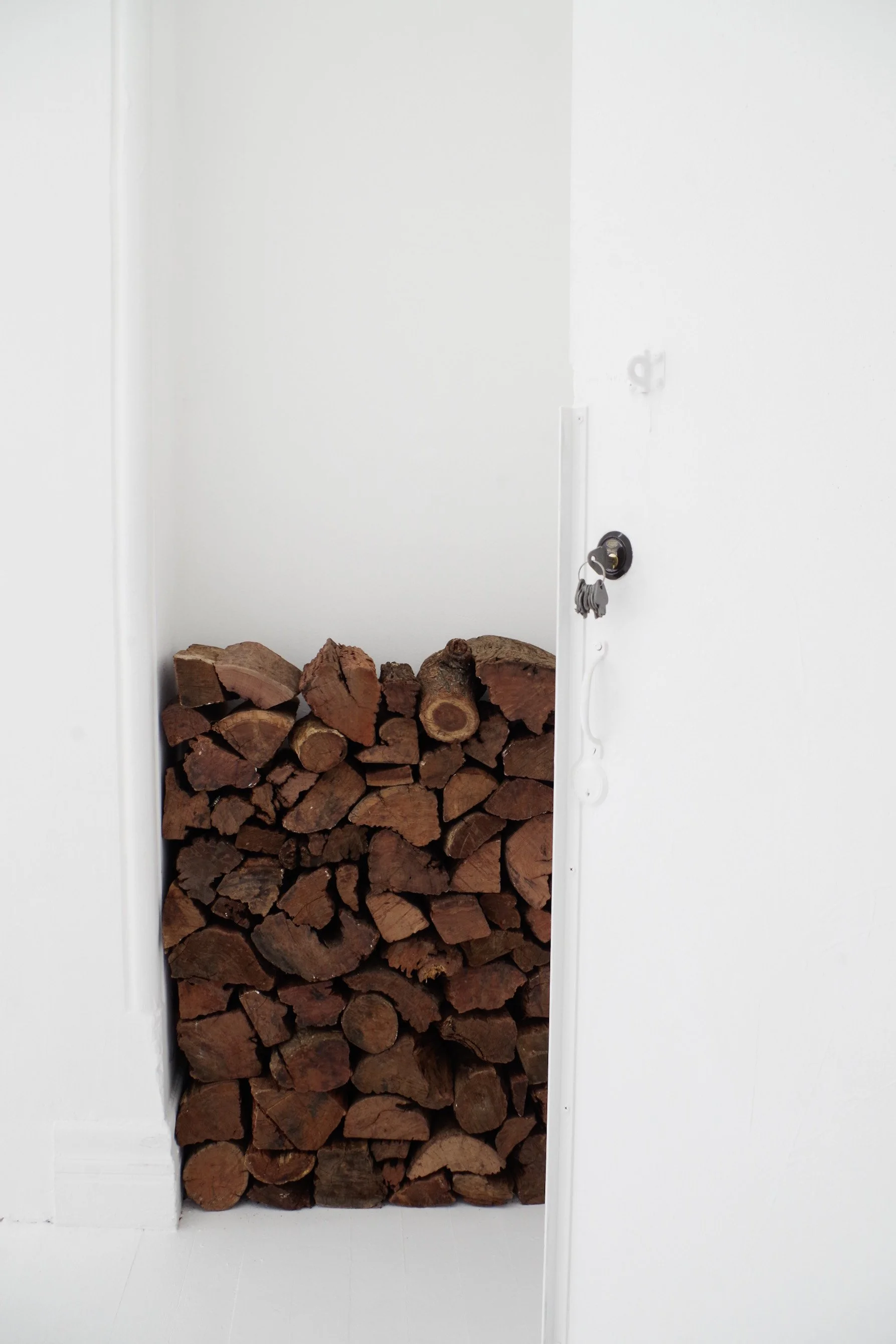 Stack of firewood logs neatly arranged in a recessed area within a white wall, next to a door with a hanging keychain.