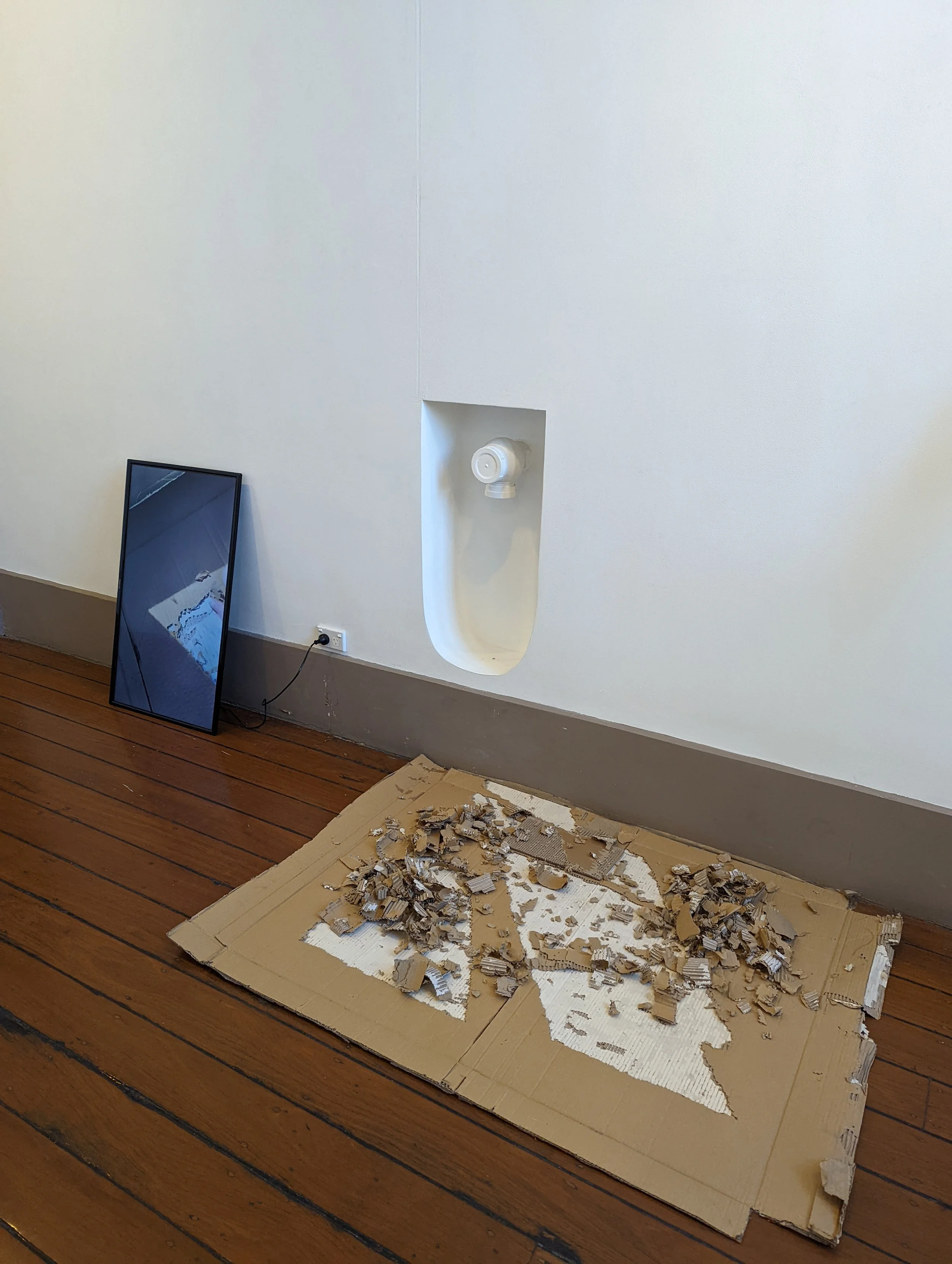 A disassembled mirror leaning against a white wall on a wooden floor with debris and torn cardboard on the ground nearby.