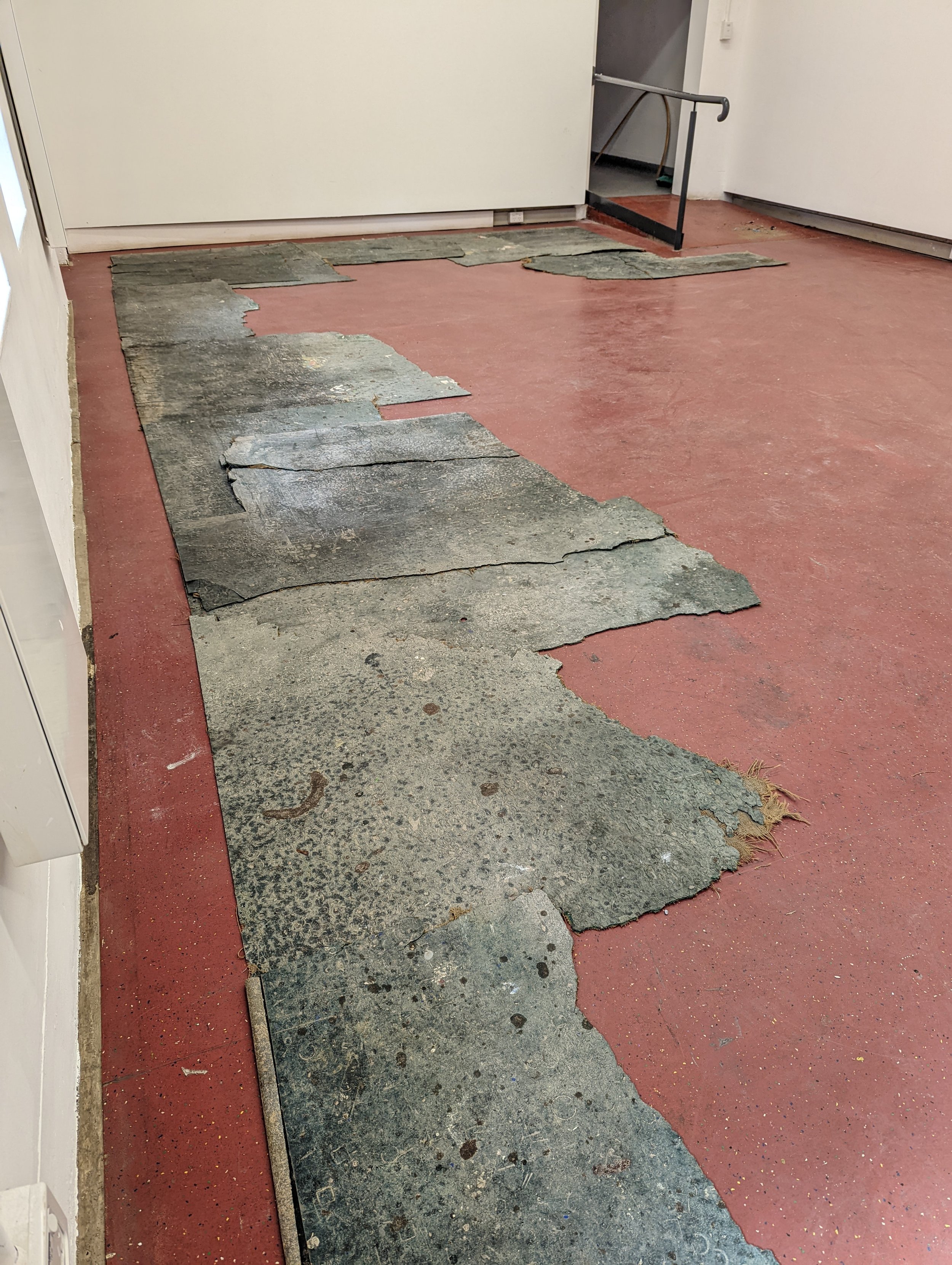 Floor with old peeling black vinyl tiles, some in broken pieces, on a red concrete surface inside a building near a white wall and a staircase with a black handrail.