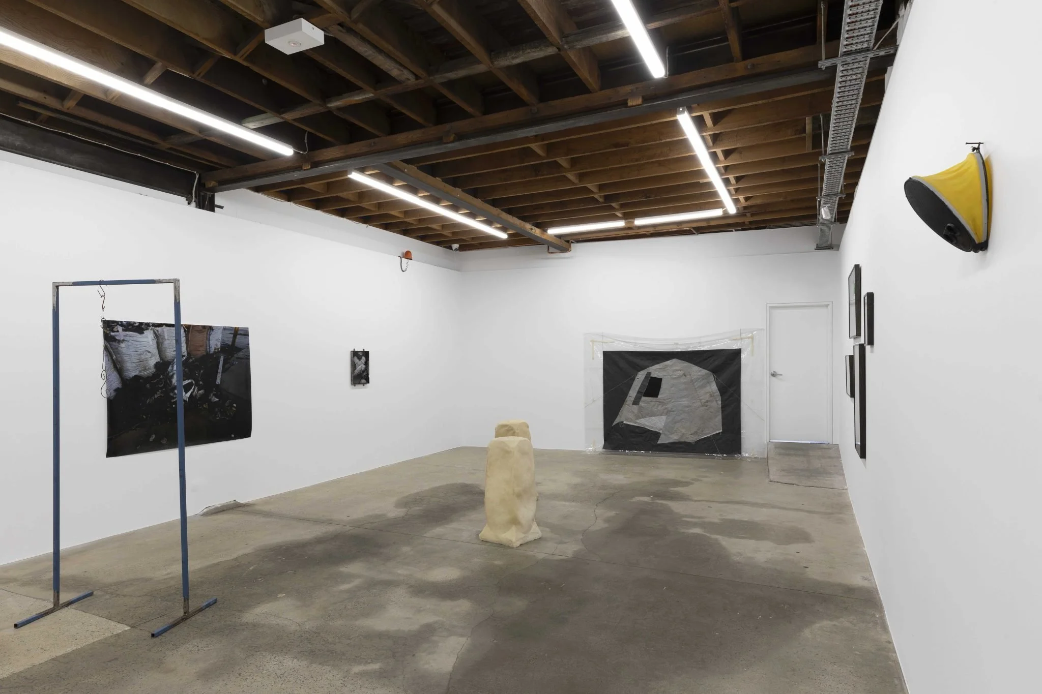 An art gallery with white walls, featuring abstract artworks, a yellow wall-mounted speaker, and installation pieces including a cloth-covered frame and a stone sculpture, under a wooden ceiling with fluorescent lighting.