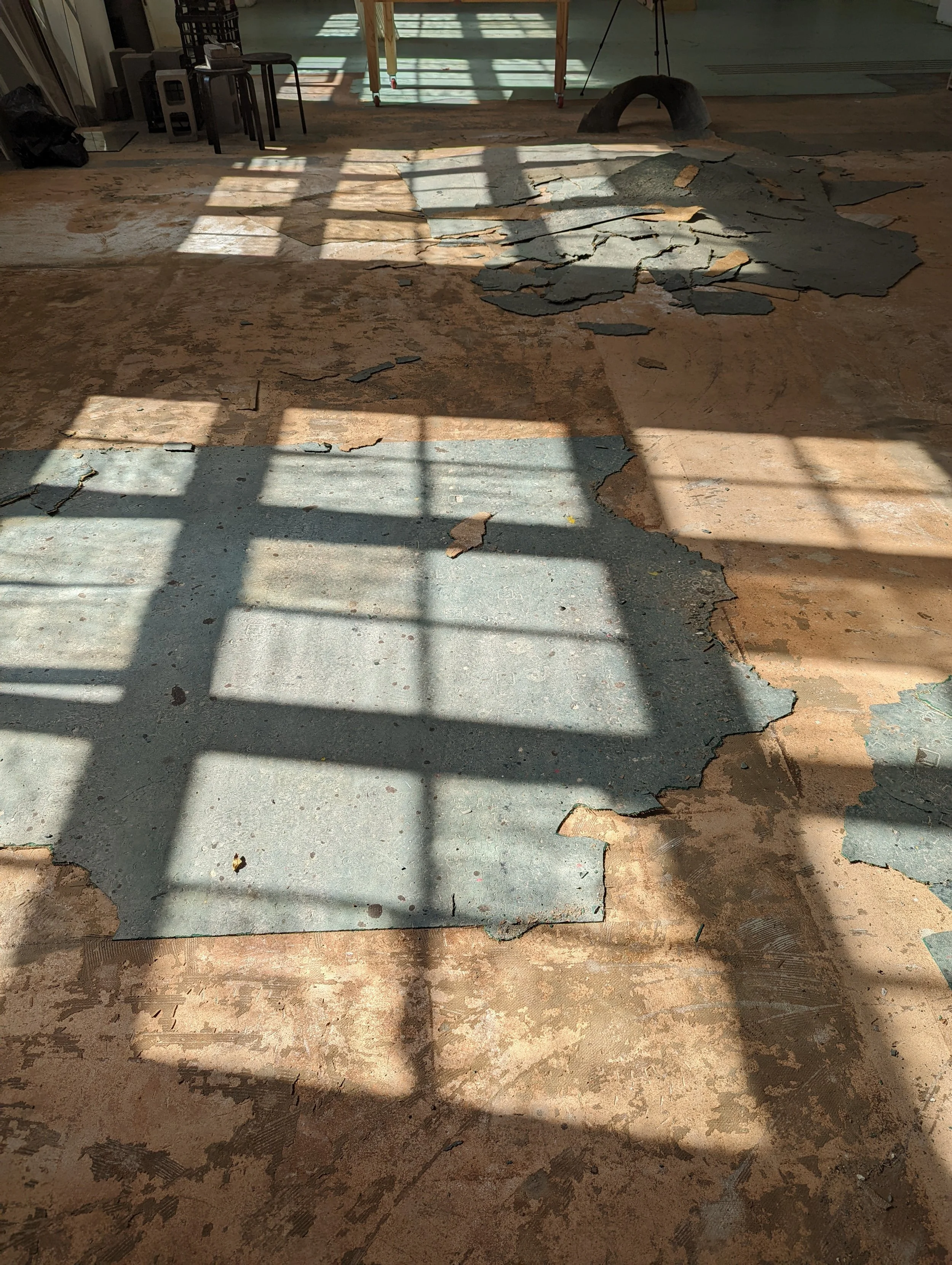 A floor with peeling paint and cracks, shadows cast from a grid structure, and some scattered debris. In the background, there are chairs, a black bag, and a table with a wooden frame and wheels.