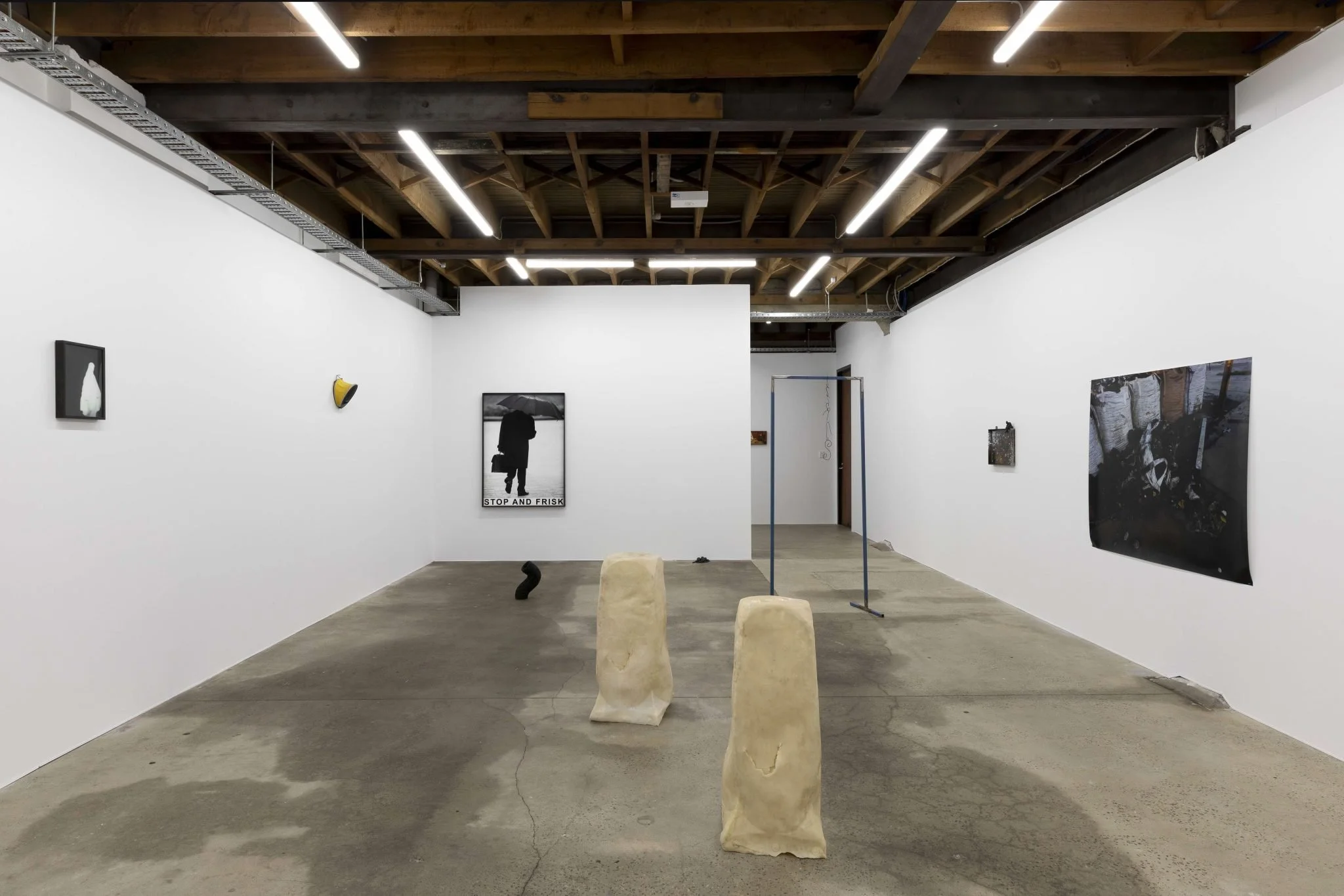 An art gallery with white walls displaying various artworks, including photographs and mixed media pieces. The gallery has a wooden ceiling with exposed beams and fluorescent lights. In the foreground, there are four sculptural stools made of light-c