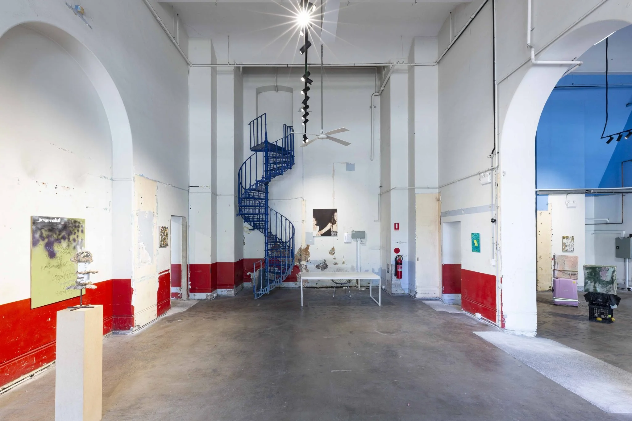 Interior of an art gallery with a spiral staircase, white walls with peeling paint, artwork on display, and a concrete floor.