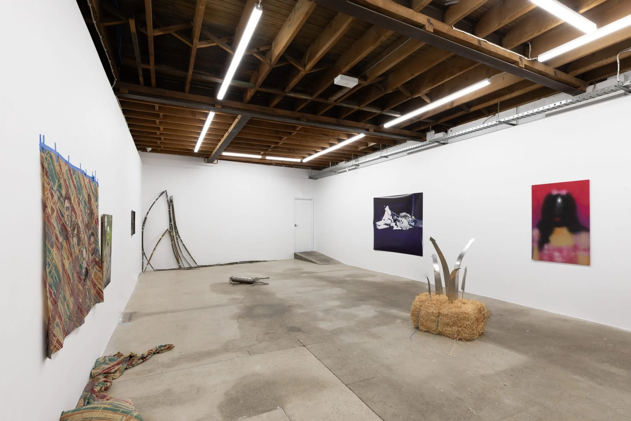 An art gallery with white walls, wooden ceiling beams, LED lights, and various framed artworks on display. A metal sculpture with curved and angular elements stands on a straw bale in the foreground. A door is visible at the back of the gallery.