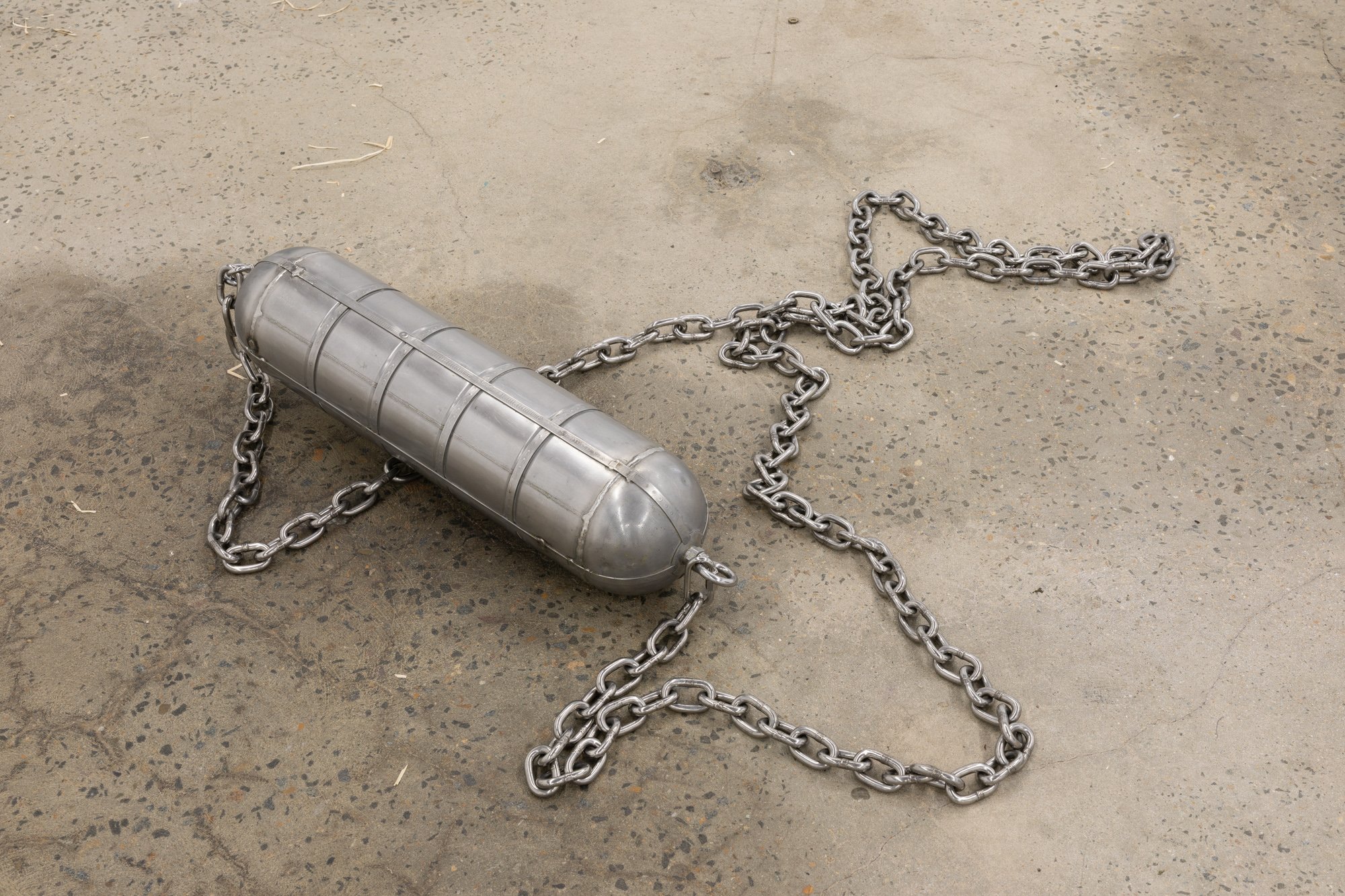 A metal bomb-shaped object lying on a concrete ground, entangled with thick silver chains.