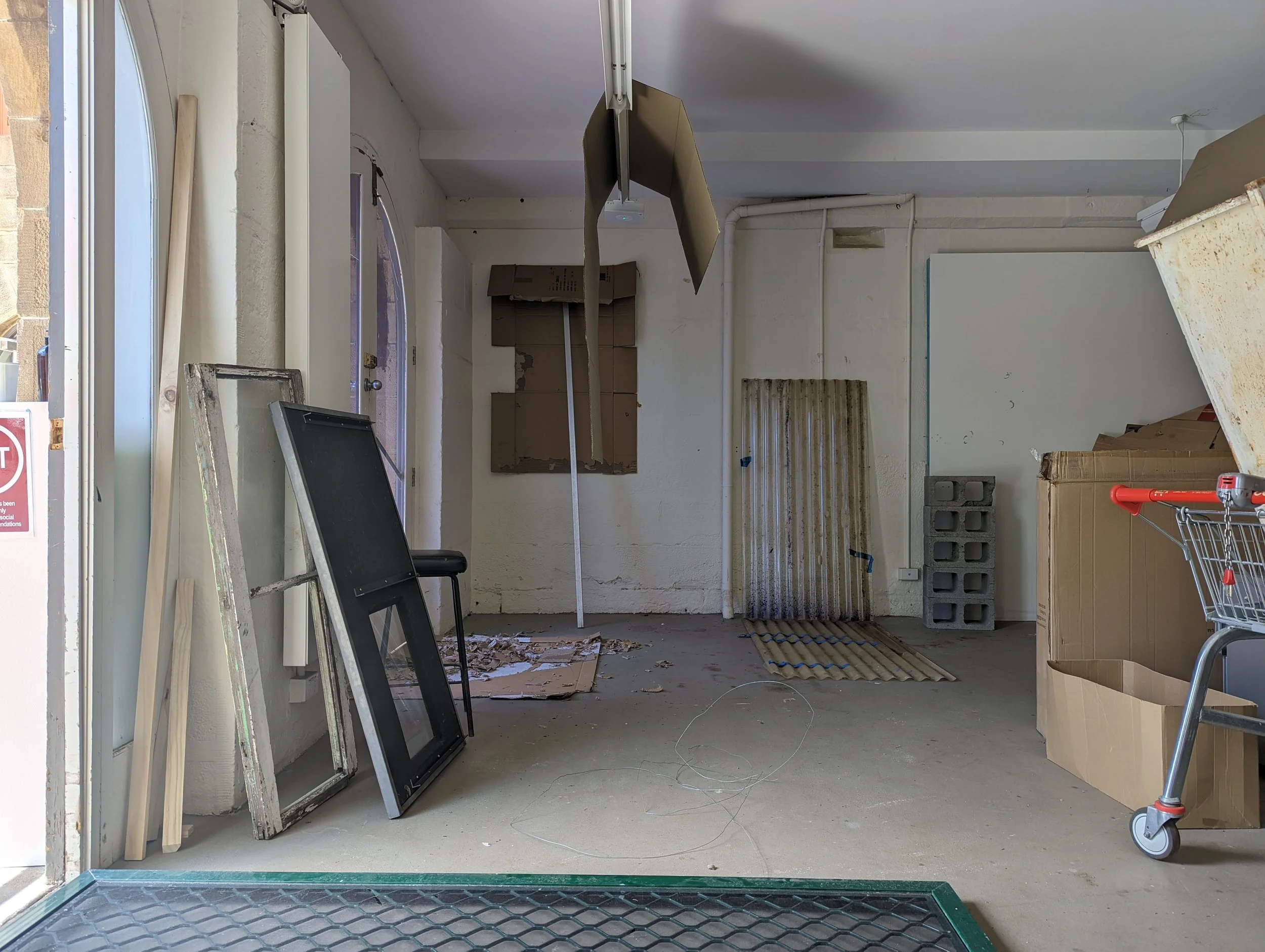 An interior view of a cluttered room or garage under renovation with construction materials, cardboard boxes, a cart, and disorganized items.
