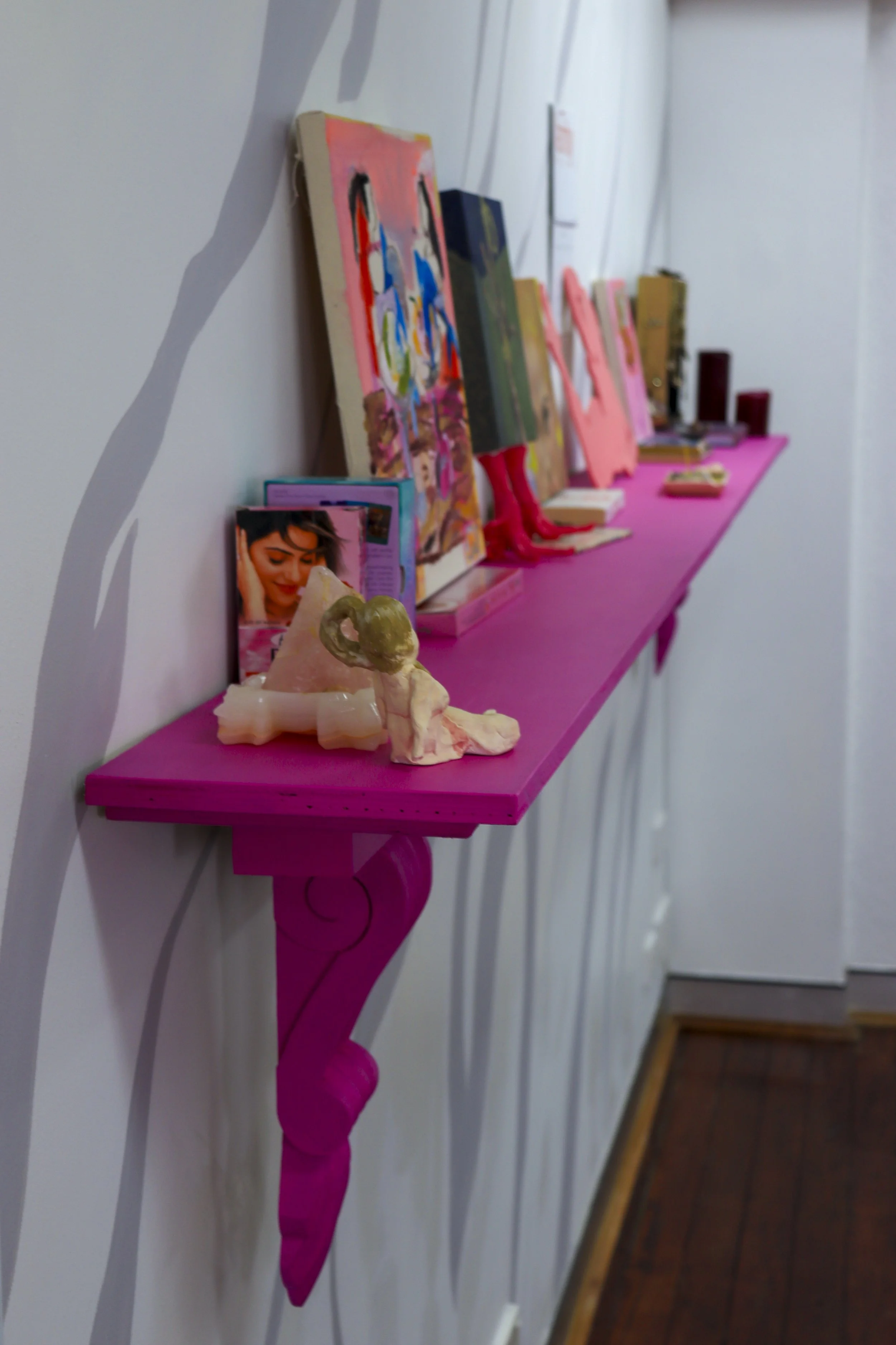 A row of small pink shelves attached to a white wall, each holding various paintings and knickknacks, with a pink table below displaying decorative items.