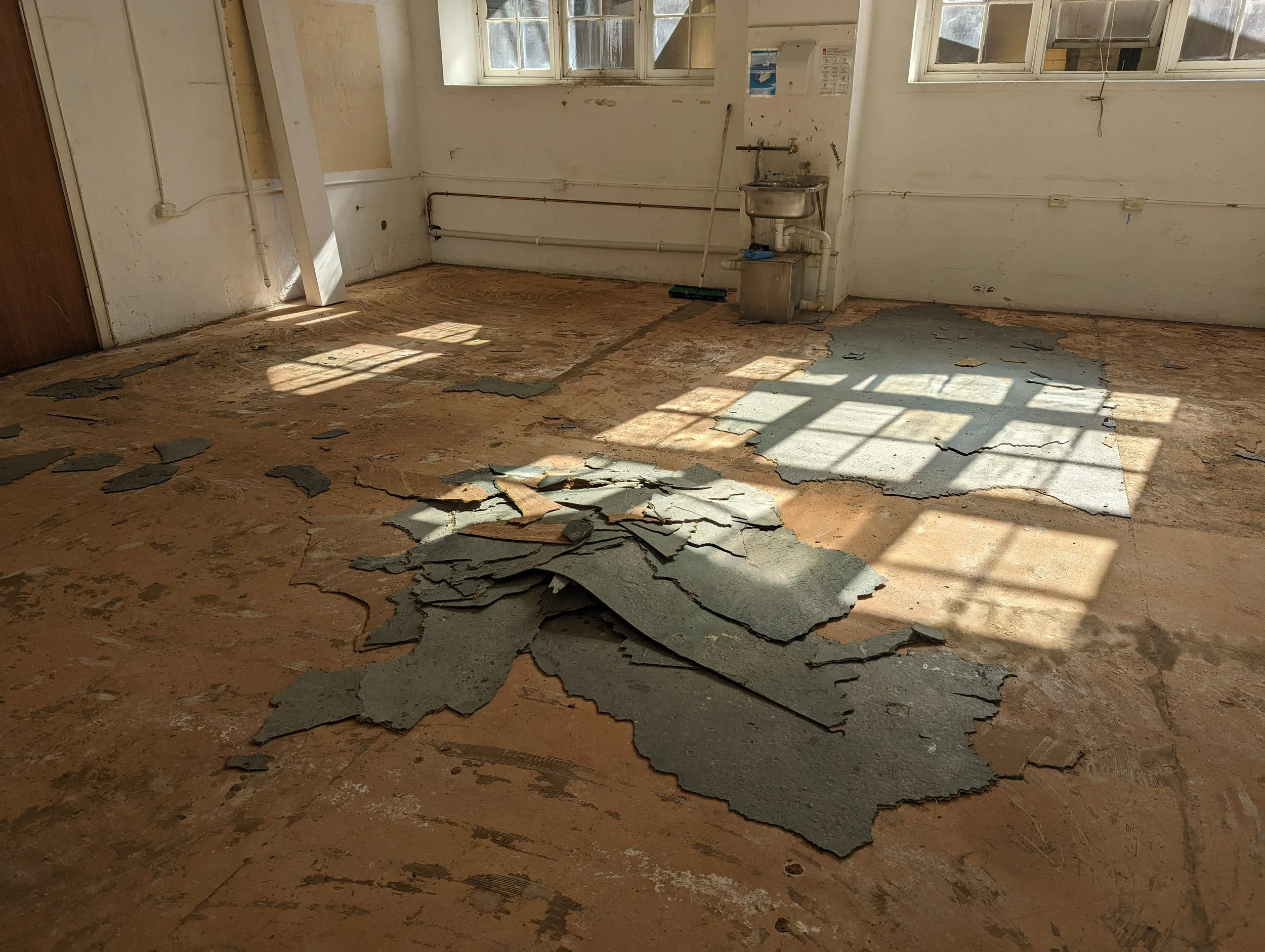 Empty room with peeling flooring, exposed pipes, and windows with sunlight casting shadows.