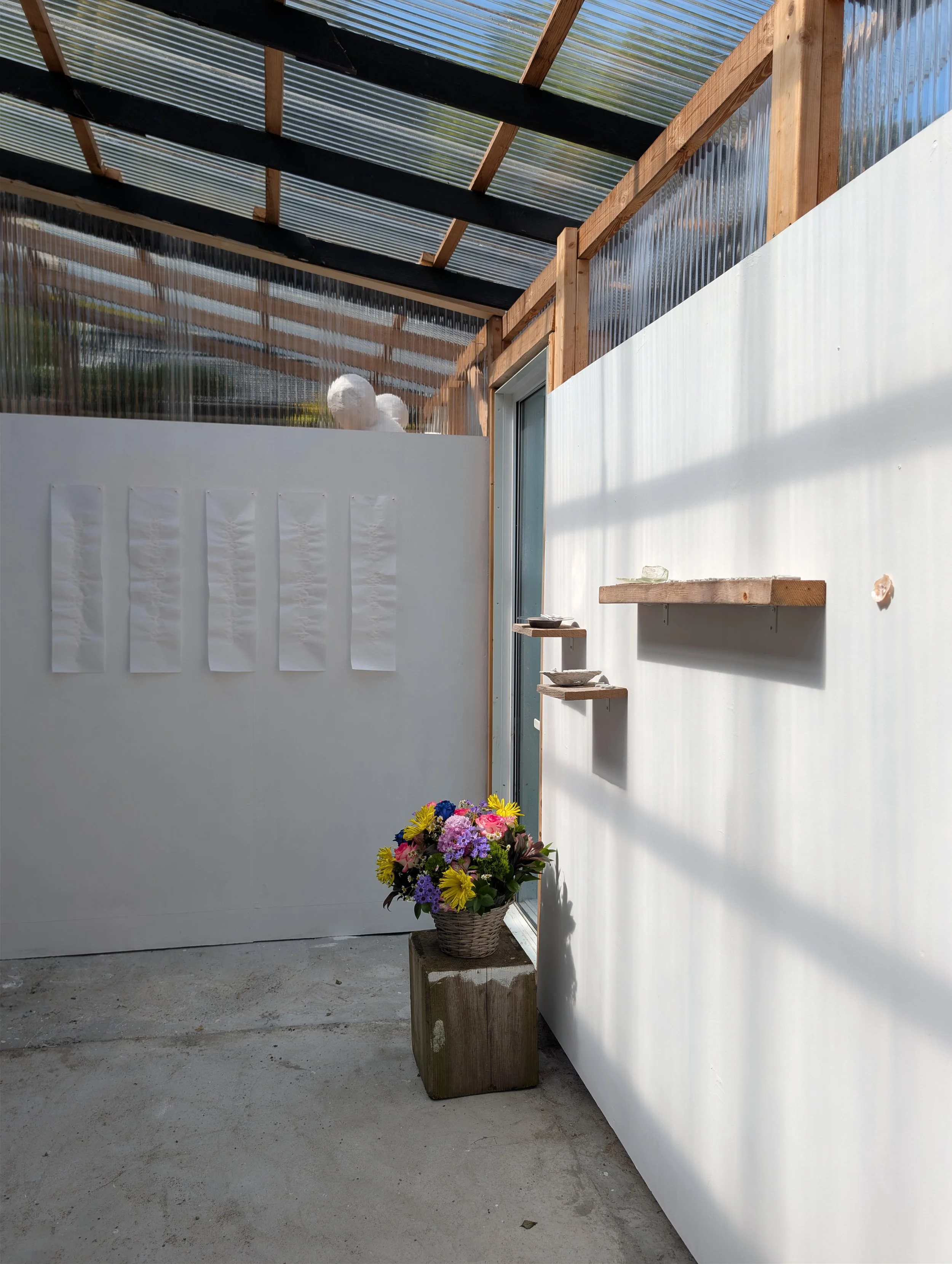 An outdoor patio or garden area with a flower arrangement basket placed on a wooden block, a white wall with hanging paper decorations on the left, a partly open door with a small wooden shelf on the right, and a transparent roof supported by wooden 