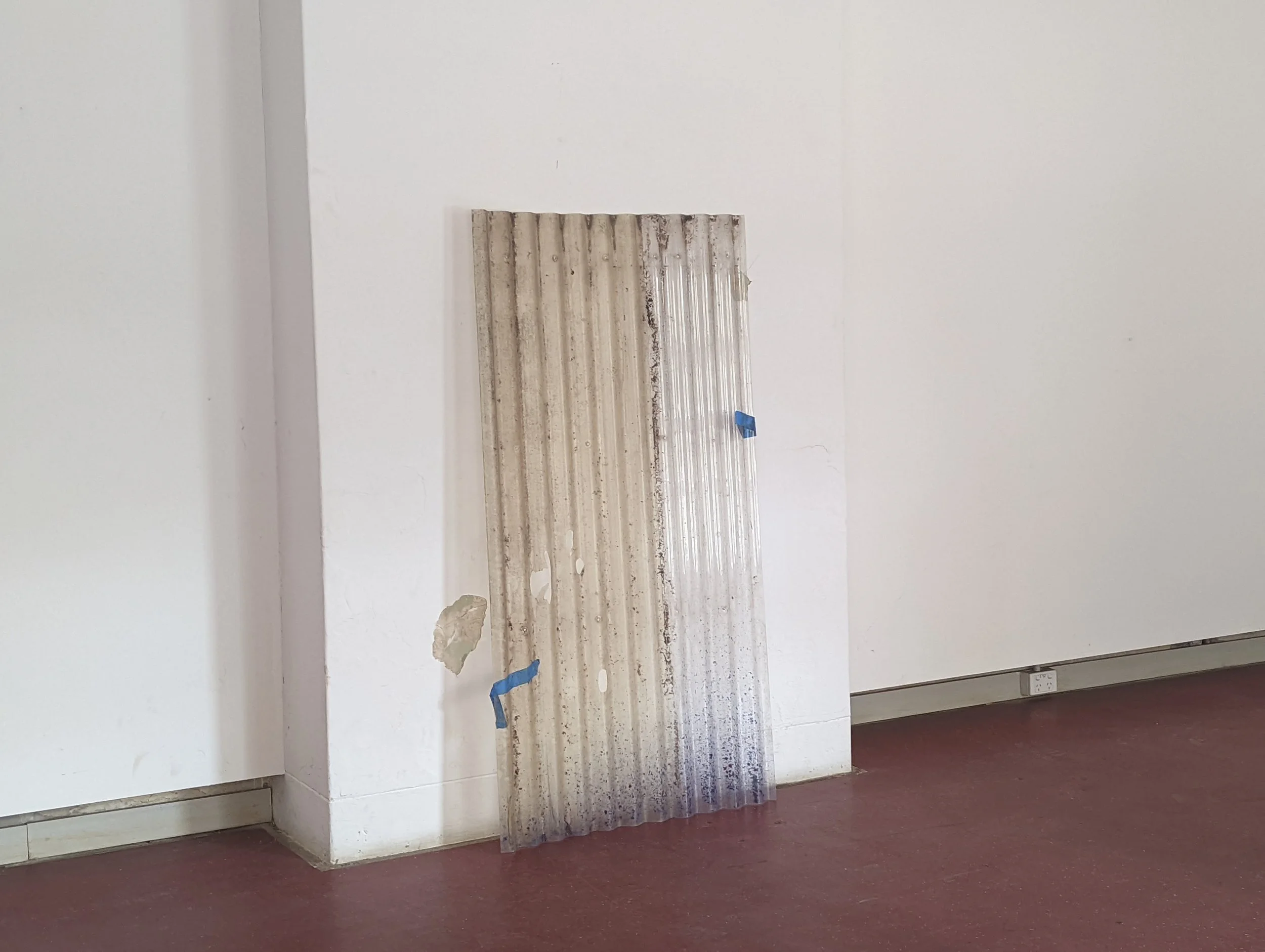 A beige and gray striped piece of corrugated metal leaning against a white wall in a room under renovation with a cut-out section and some debris on the floor.