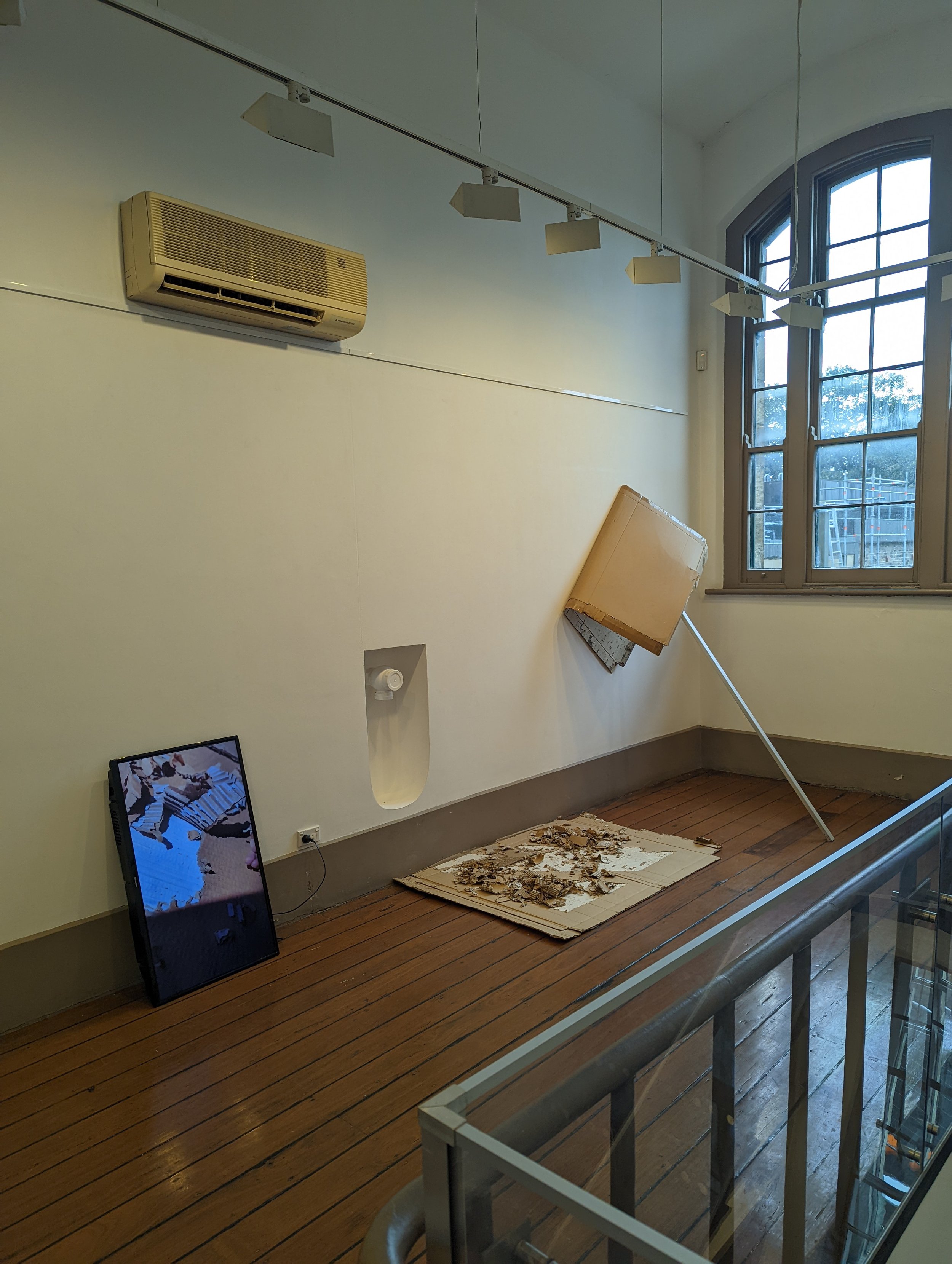 Interior of a building under renovation with large windows, a wall-mounted air conditioning unit, a pole leaning against the wall, debris on the floor, and a fallen cardboard box.