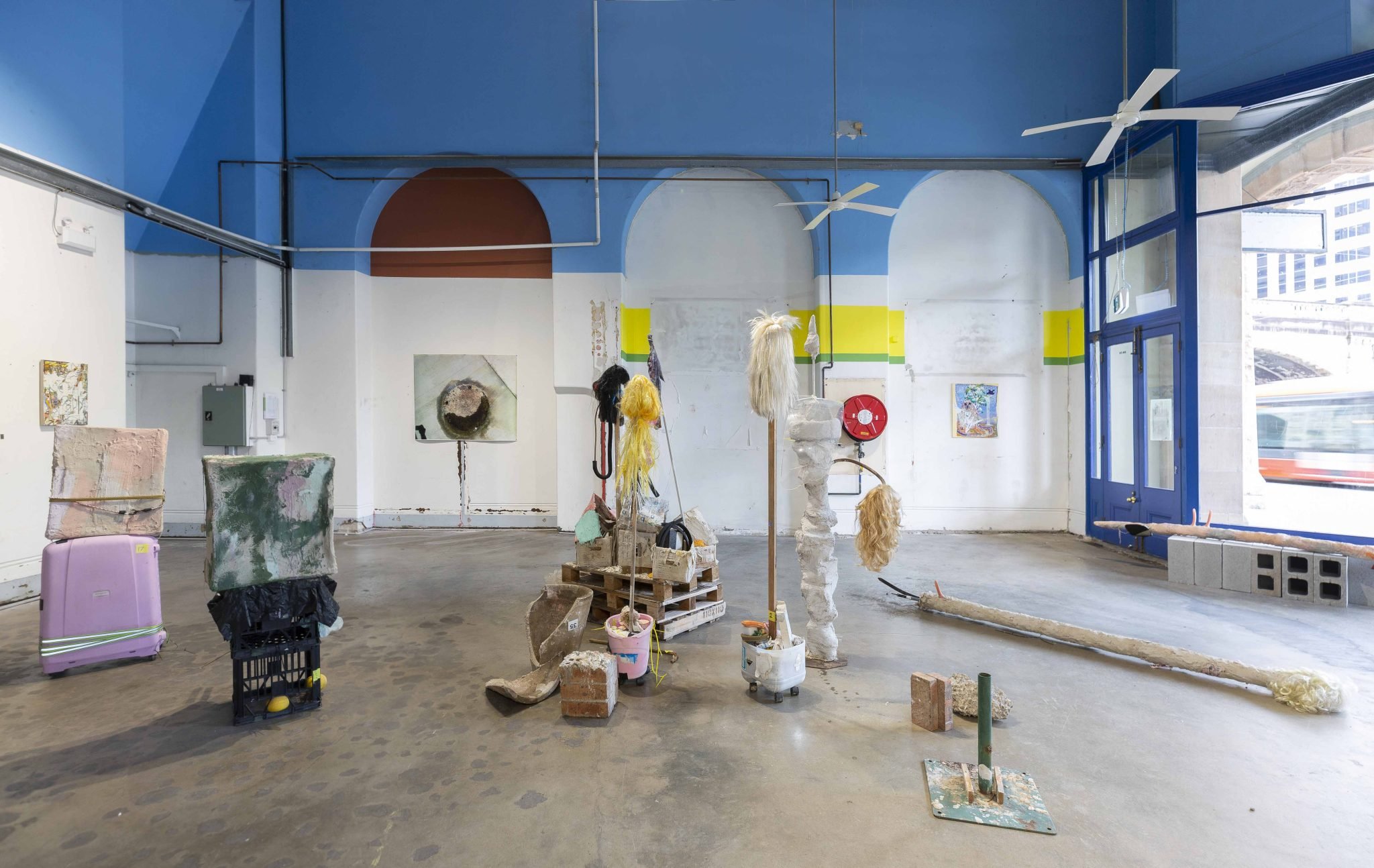 Art studio with colorful painted walls, sculptures, and art supplies, including hangings of wigs, boxes, and various art pieces, visible through a large window and blue door.