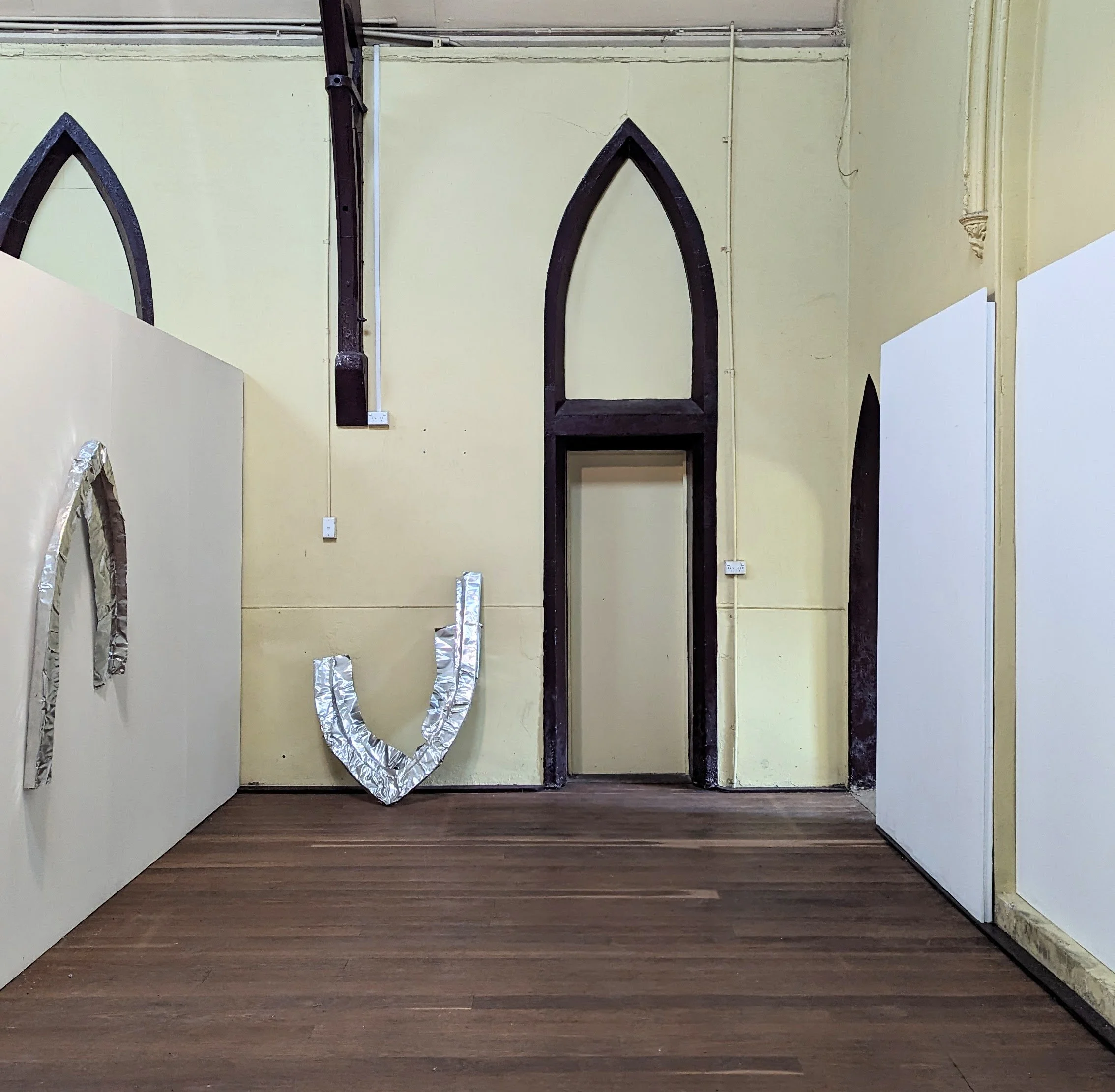 Interior of a room under construction or renovation with black gothic-style window frames, yellow walls, and a wood floor. There is foil-wrapped material and electrical outlets on the walls, and exposed pipes and wires at the ceiling.