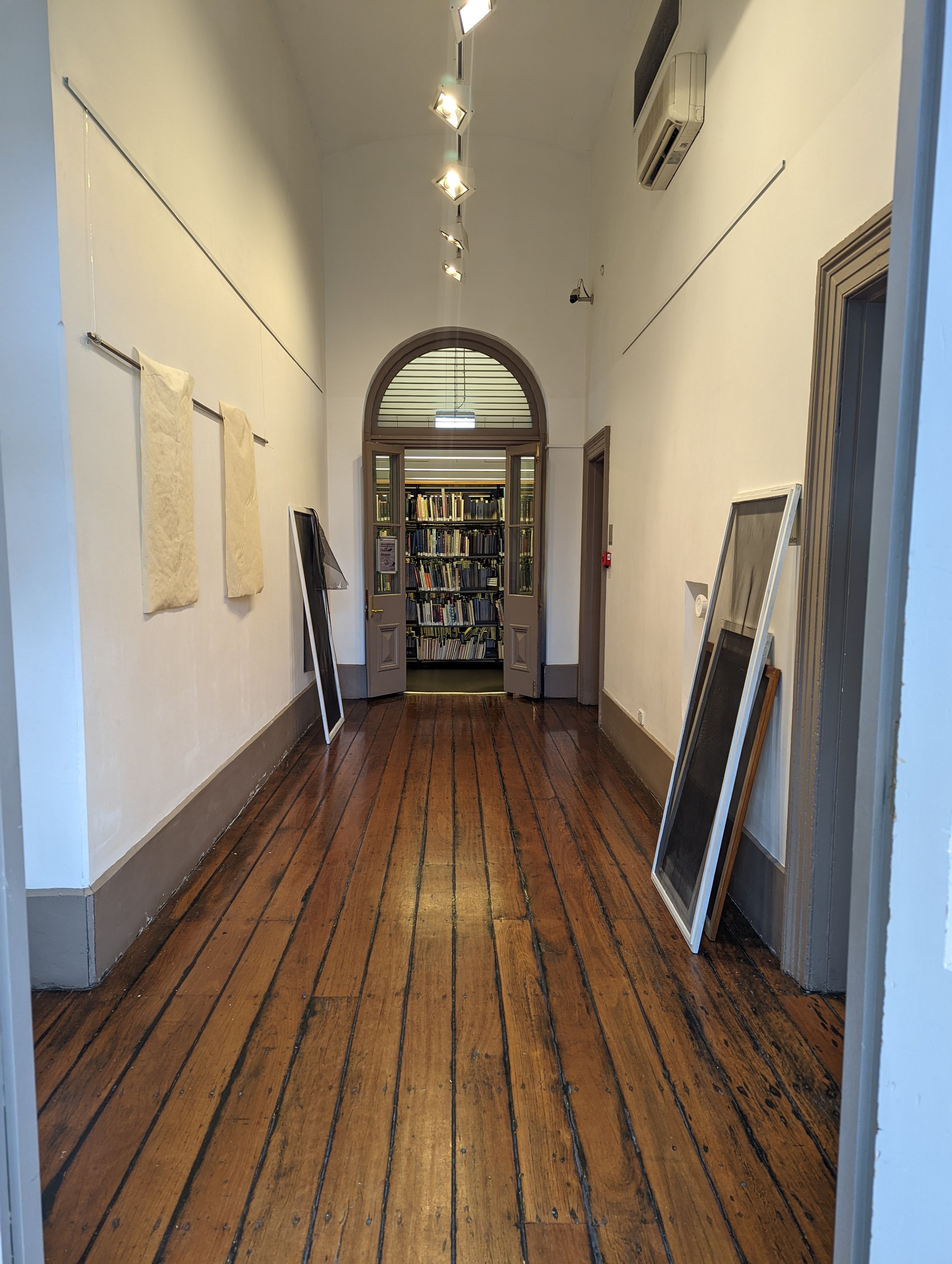 A hallway with wooden floors, white walls, and framed artworks leaning against the wall, with a set of open double doors leading to a bookcase filled with books.