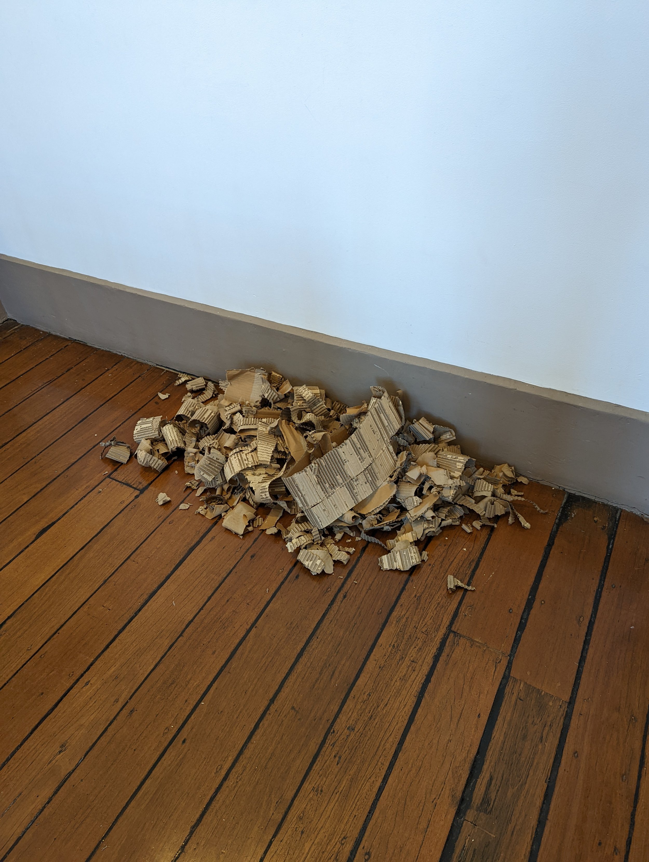 A pile of shredded cardboard on a wooden floor next to a blue wall with a grey baseboard.