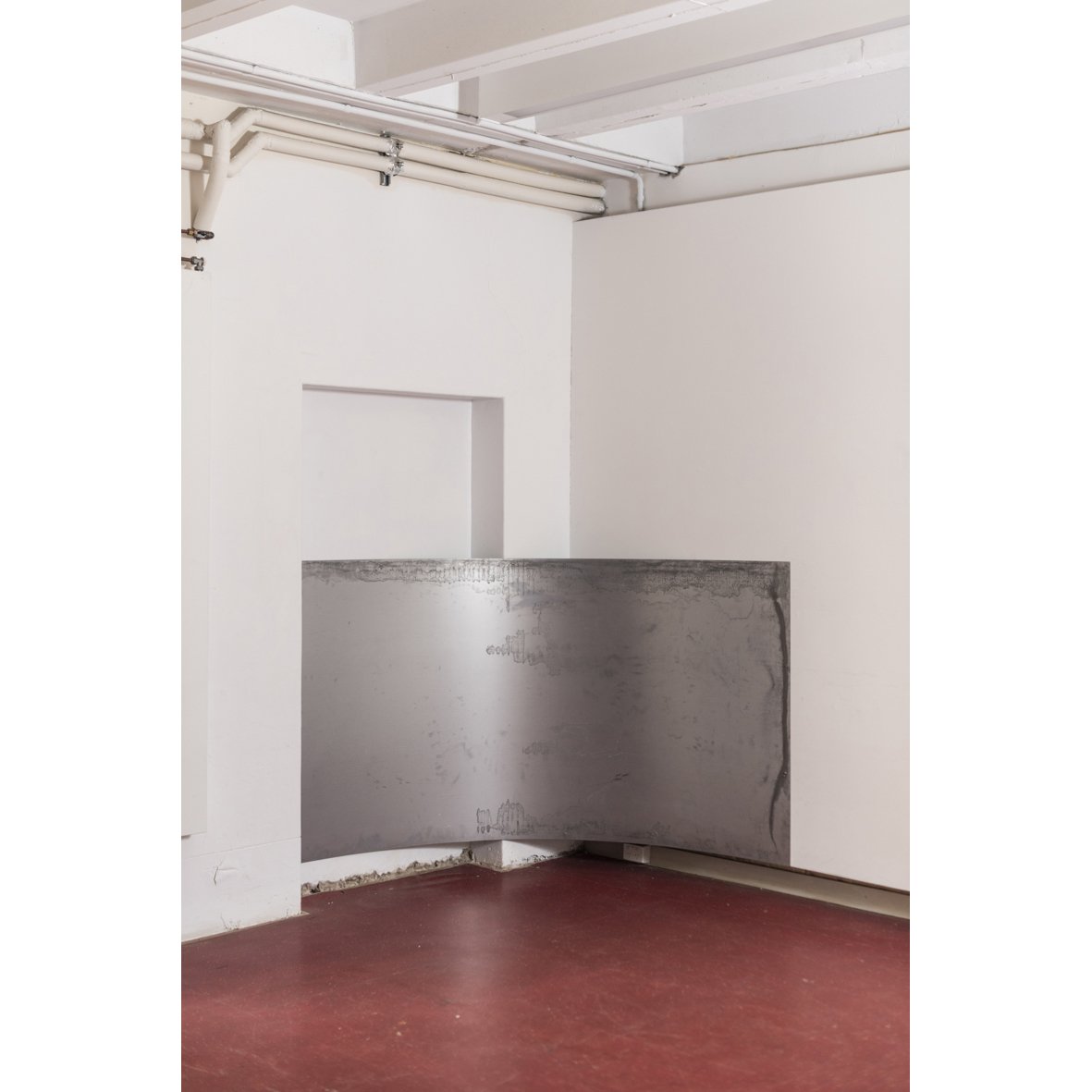 Empty room with white walls, exposed pipes on ceiling, concrete barrier, and red floor.