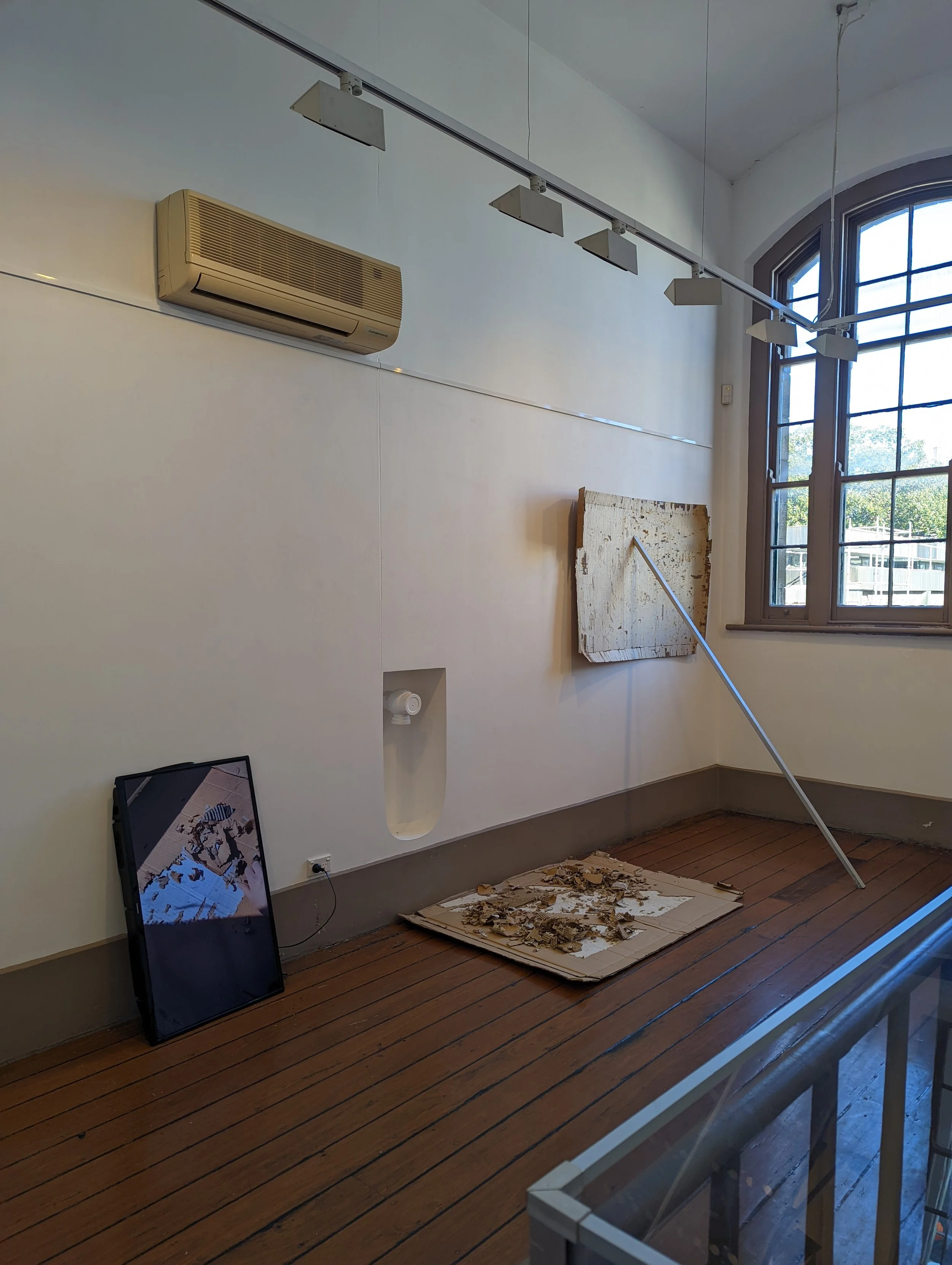 An interior space with a high ceiling, large arched windows, and a wooden floor. A broken piece of artwork or a sign leans against the wall, a disturbed cardboard and debris on the floor, and a framed image on the floor. A beige air conditioning unit