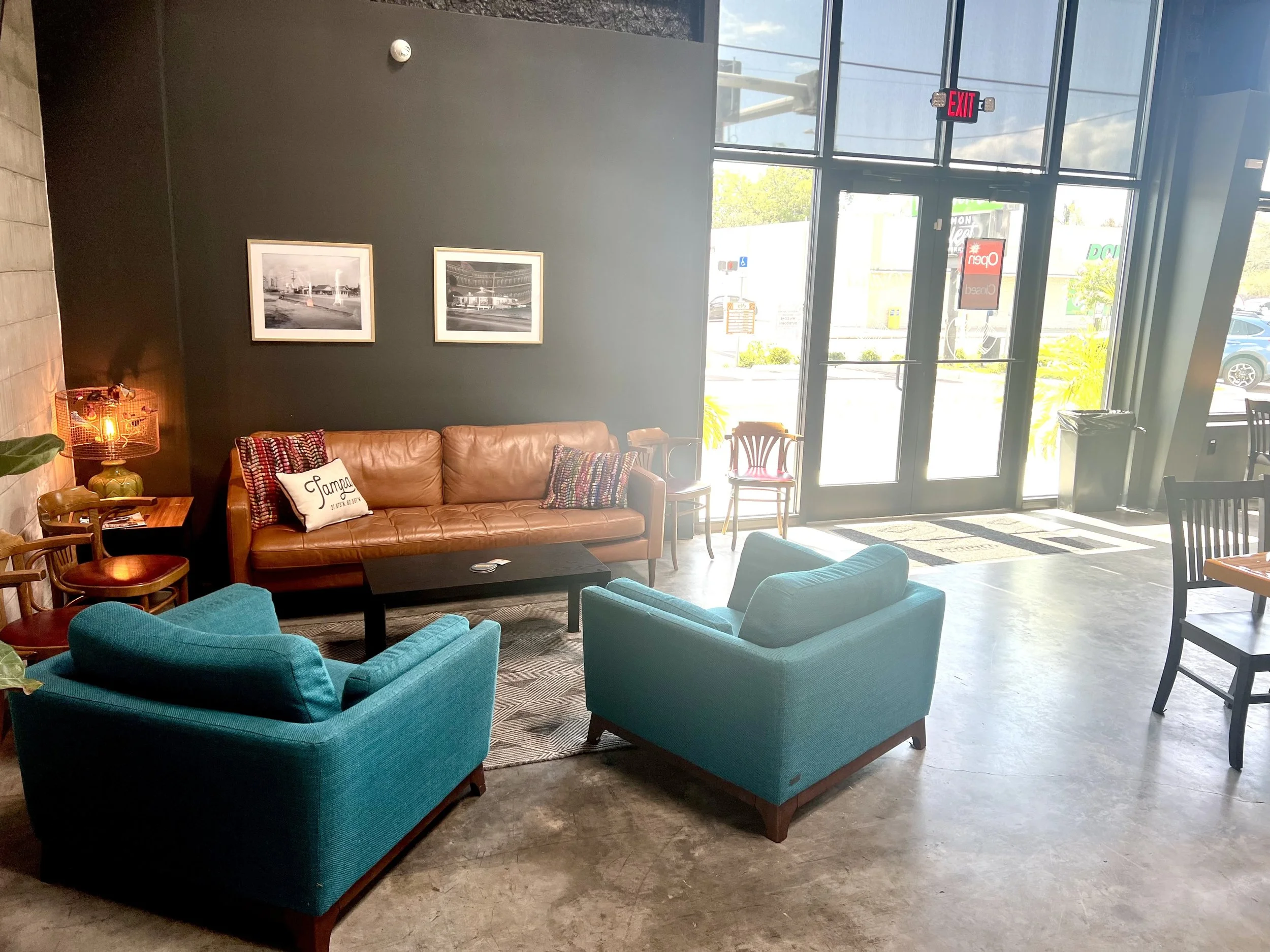 Interior of a modern waiting or lounge area with teal and brown armchairs, a leather sofa, framed black and white photos on a dark wall, and large glass windows and doors allowing sunlight inside.