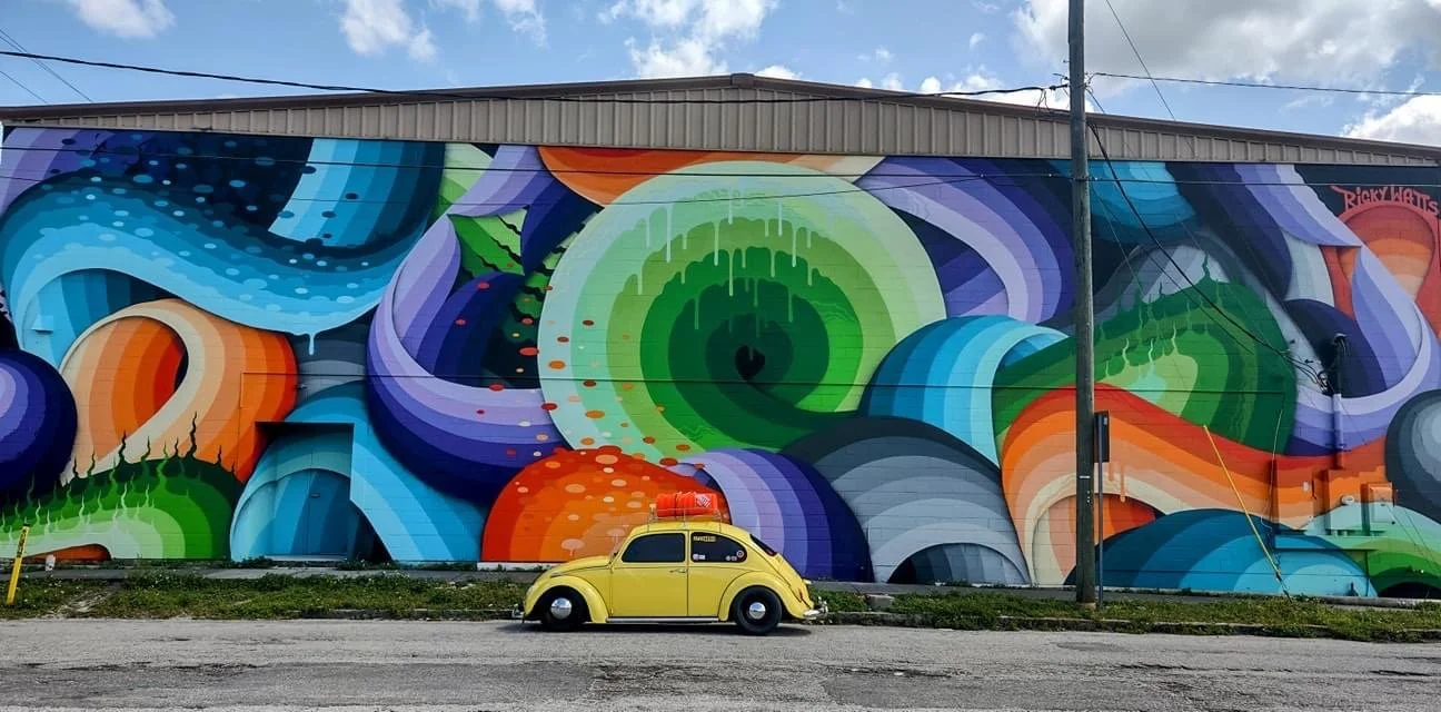 Colorful mural with abstract swirling shapes and patterns on a building wall, featuring large shapes in shades of green, blue, purple, orange, and black. A small yellow vintage car is parked in front.
