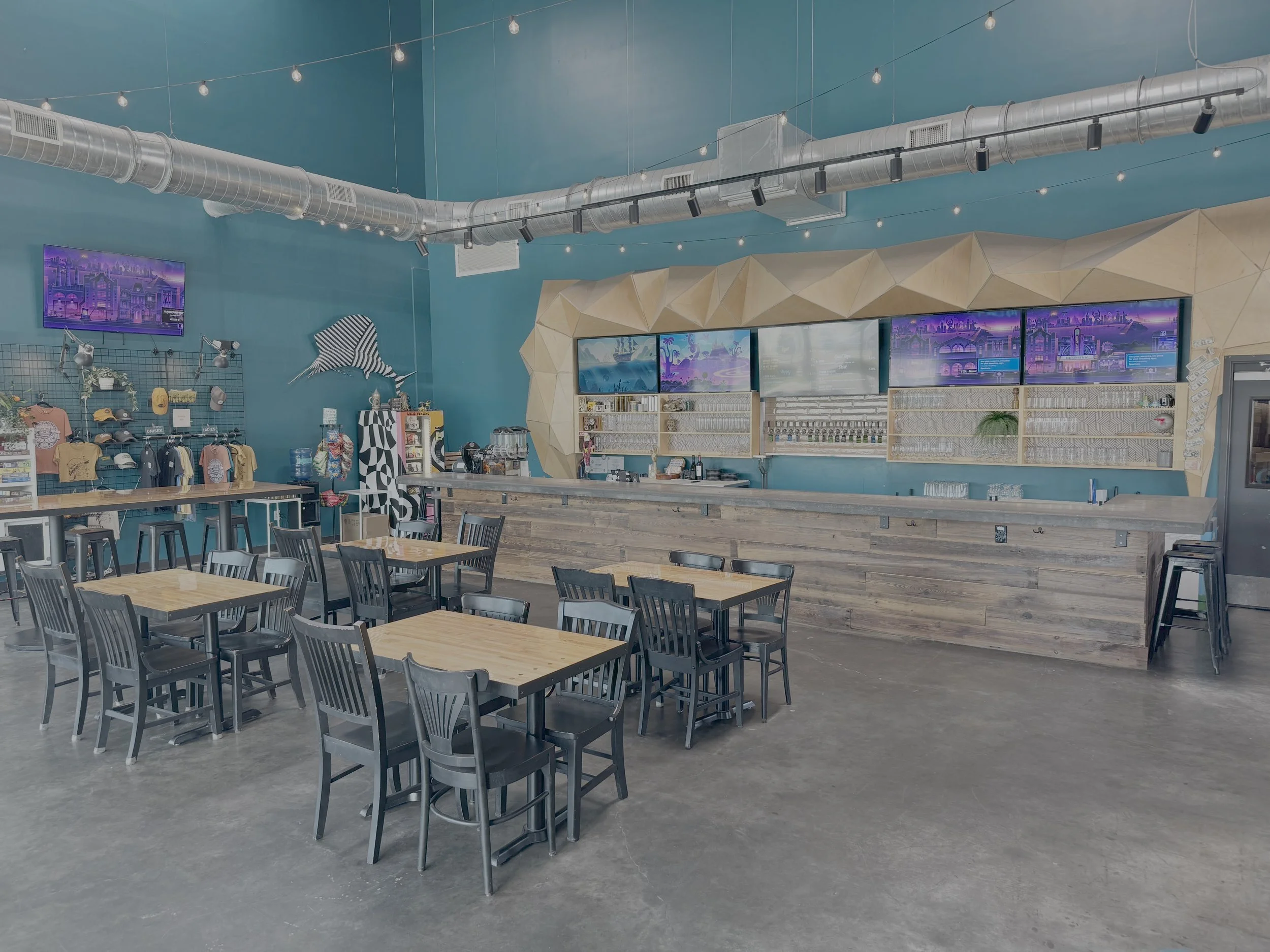 Interior of a modern cafe or restaurant with wooden tables and black chairs, ocean-themed decor, multiple flat screen monitors on the wall, teal painted walls, exposed ductwork, string lights hanging from the ceiling, and a small bar area with glasse