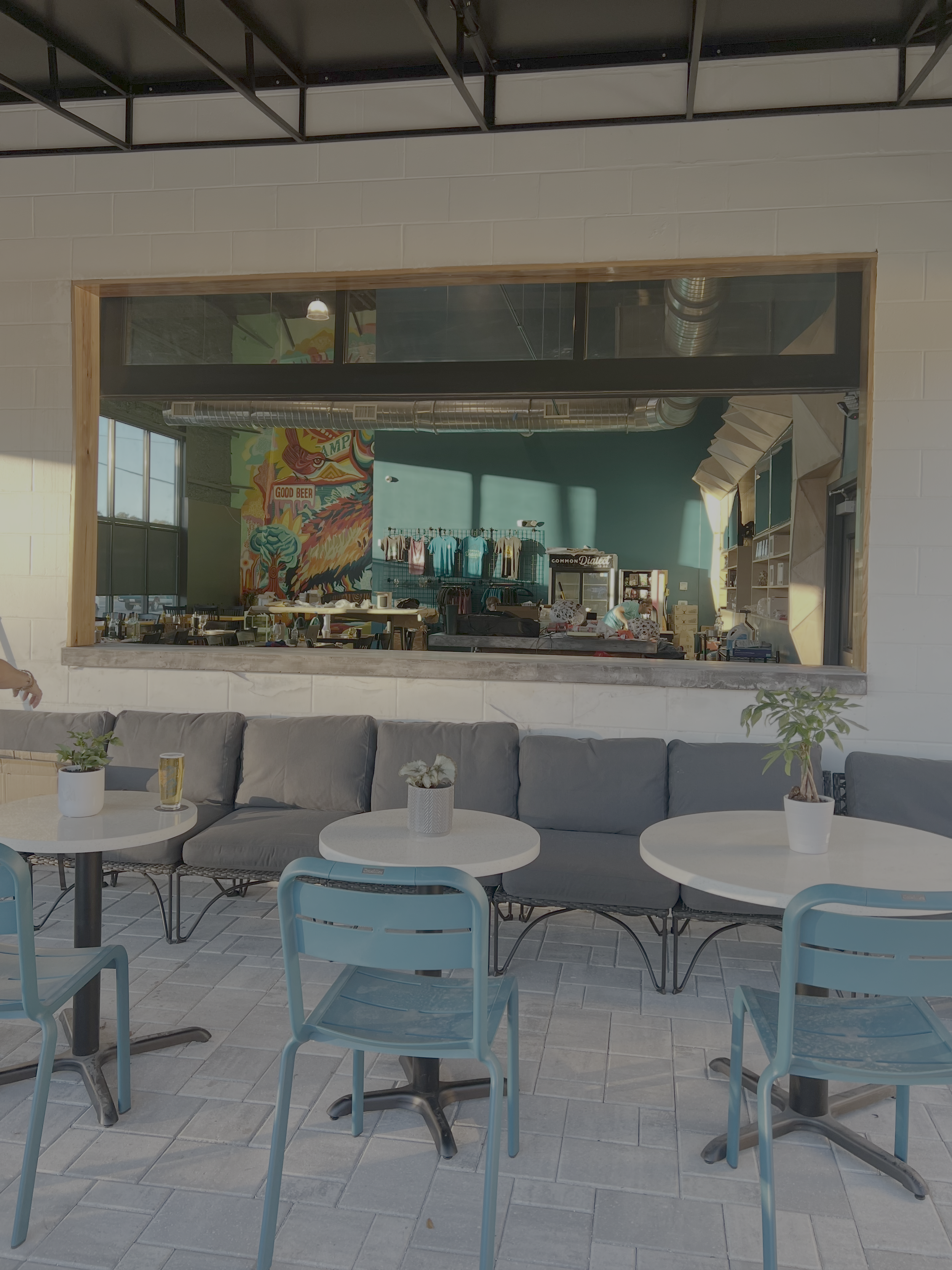 Interior of a modern cafe with tables, chairs, potted plants, a colorful mural on the wall, and a large window showing natural light.
