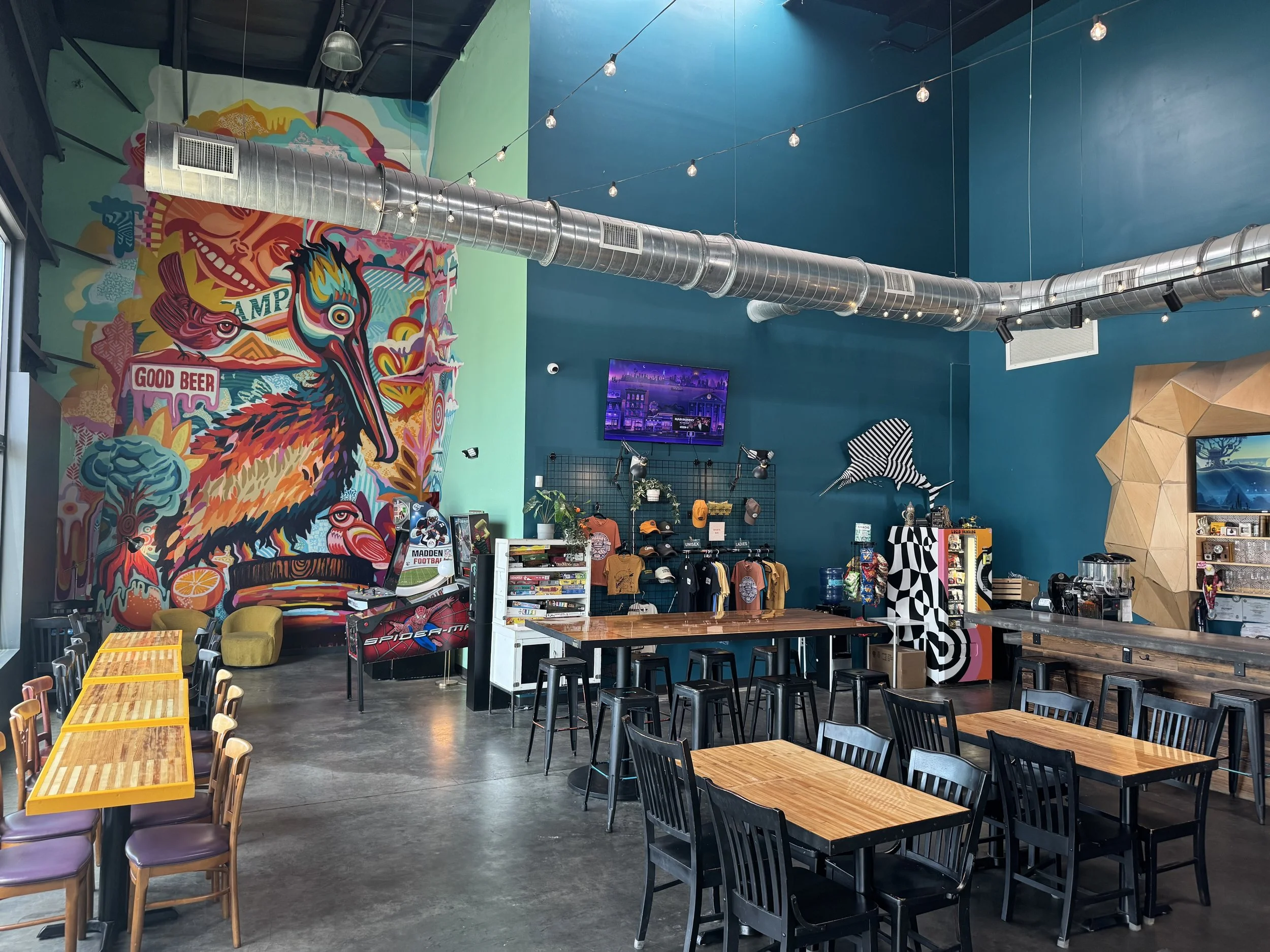 Interior of a modern cafe or restaurant with colorful graffiti art on the wall, including a dragon, a fish, and the words 'Good Beer,' with high tables, chairs, and a large ventilation duct on the ceiling. Event space Tampa
