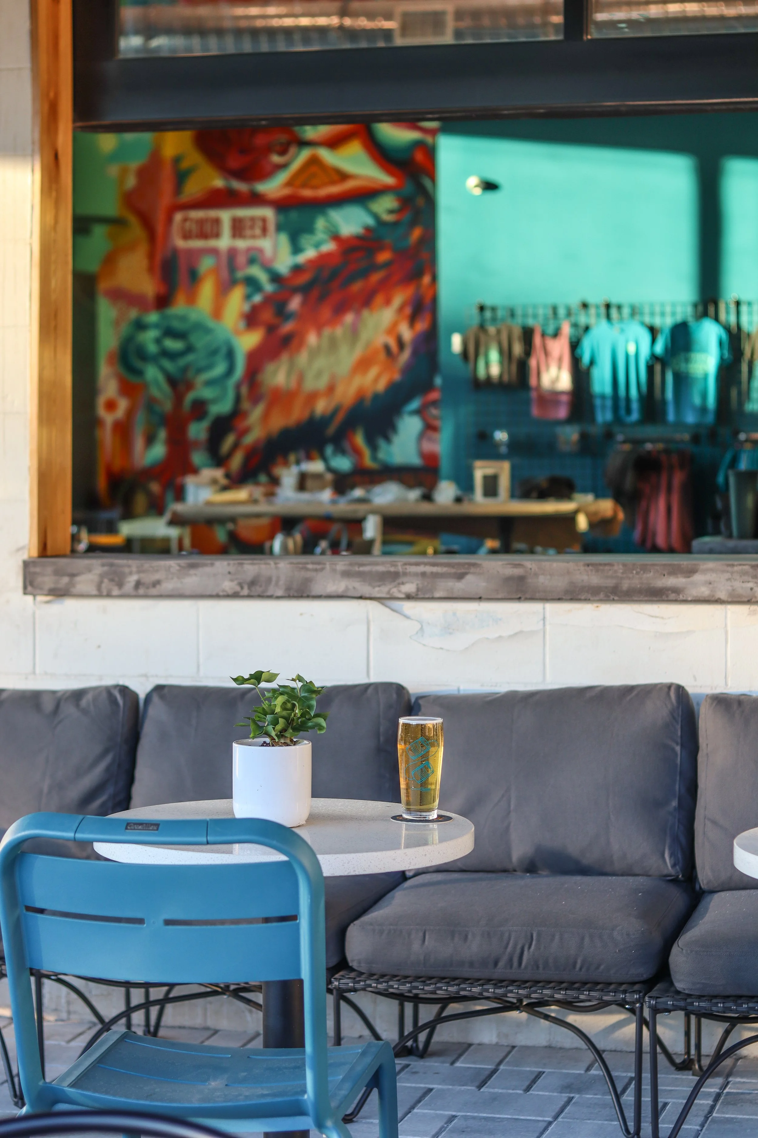 A cozy cafe scene featuring a small white round table with a potted green plant and a glass of beer, a black cushioned bench, a blue chair, and a large colorful artwork of a bird, with additional clothing items hanging on a rack inside.