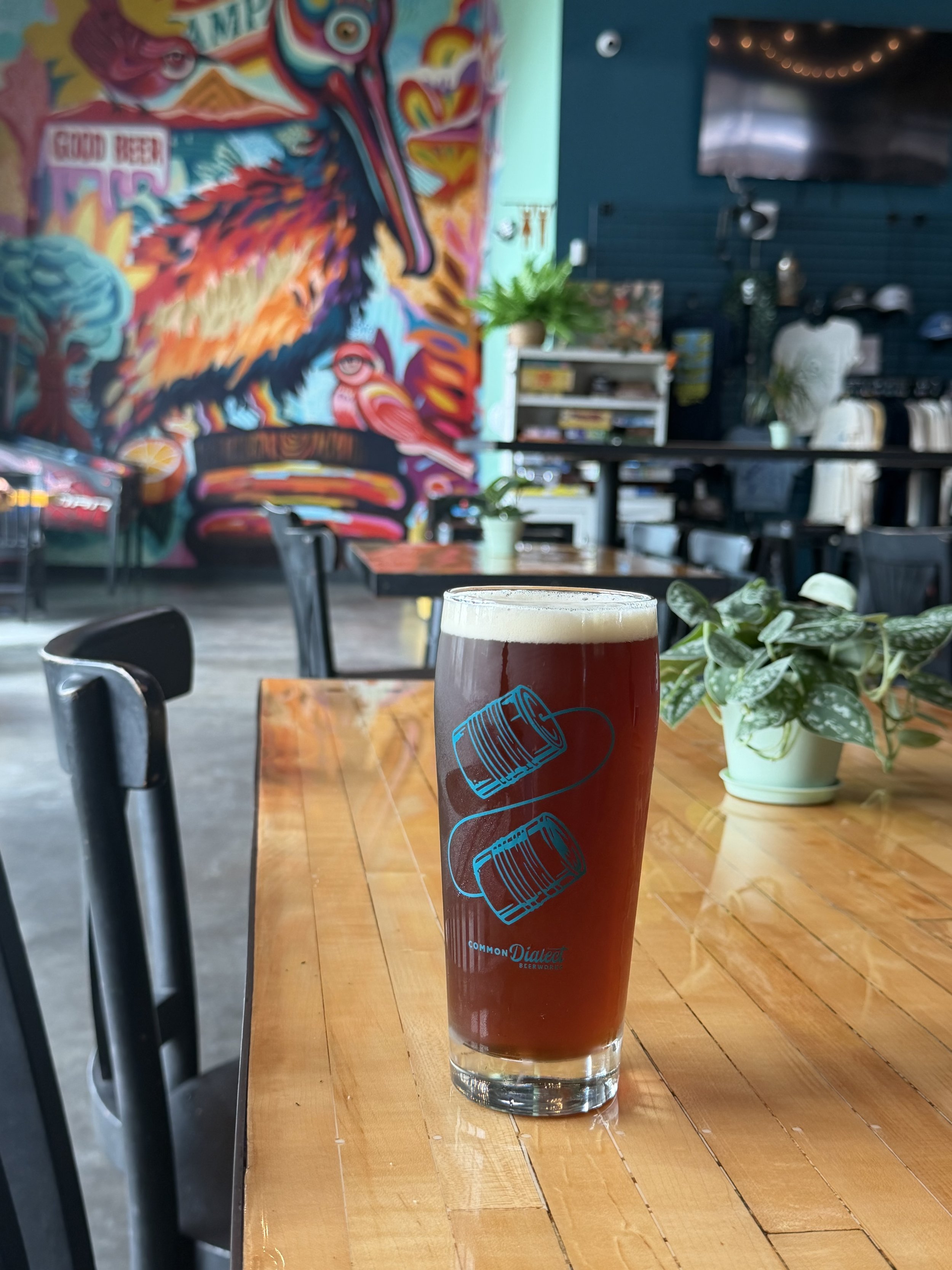A pint of beer on a wooden table with a plant in a white pot beside it in a cozy indoor setting decorated with green plants, a colorful mural on the wall, and a television mounted on a dark blue wall in the background.