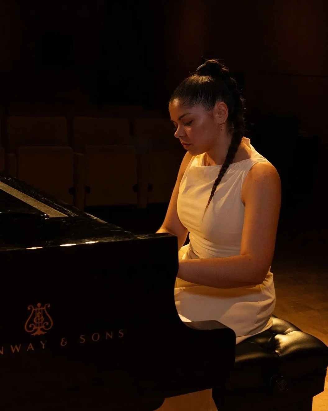 A woman with dark hair styled in a braid, wearing a sleeveless cream dress, playing a grand piano in a dimly lit room.