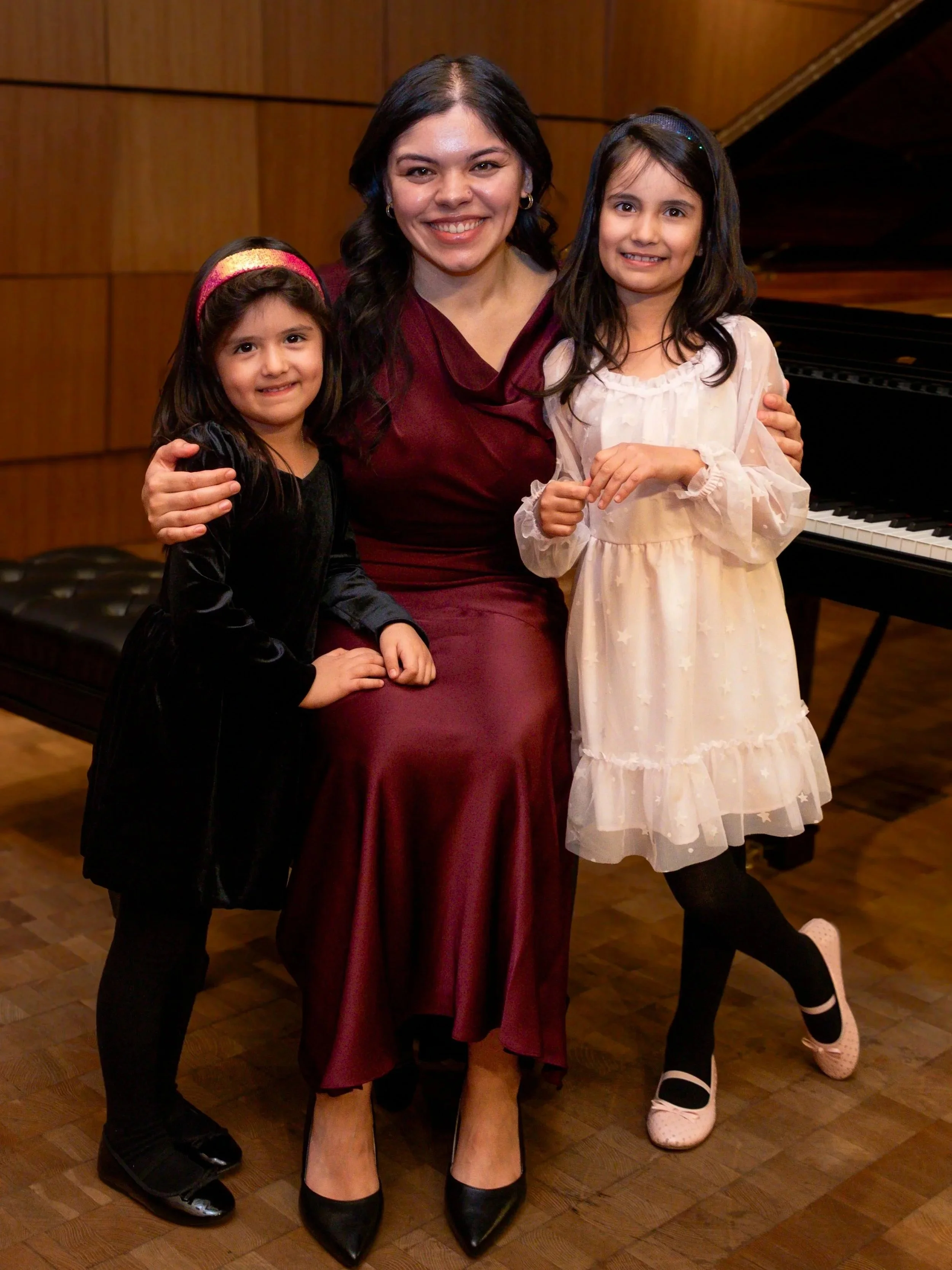 A woman in a maroon dress sitting on a piano stool, smiling. Two young girls are standing beside her, also smiling, with their arms around her. The girl on the left has a black velvet dress and pink headband, while the girl on the right wears a white dress with star patterns and ballet shoes. The setting appears to be a music or recital hall with wood-paneled walls and a grand piano in the background.