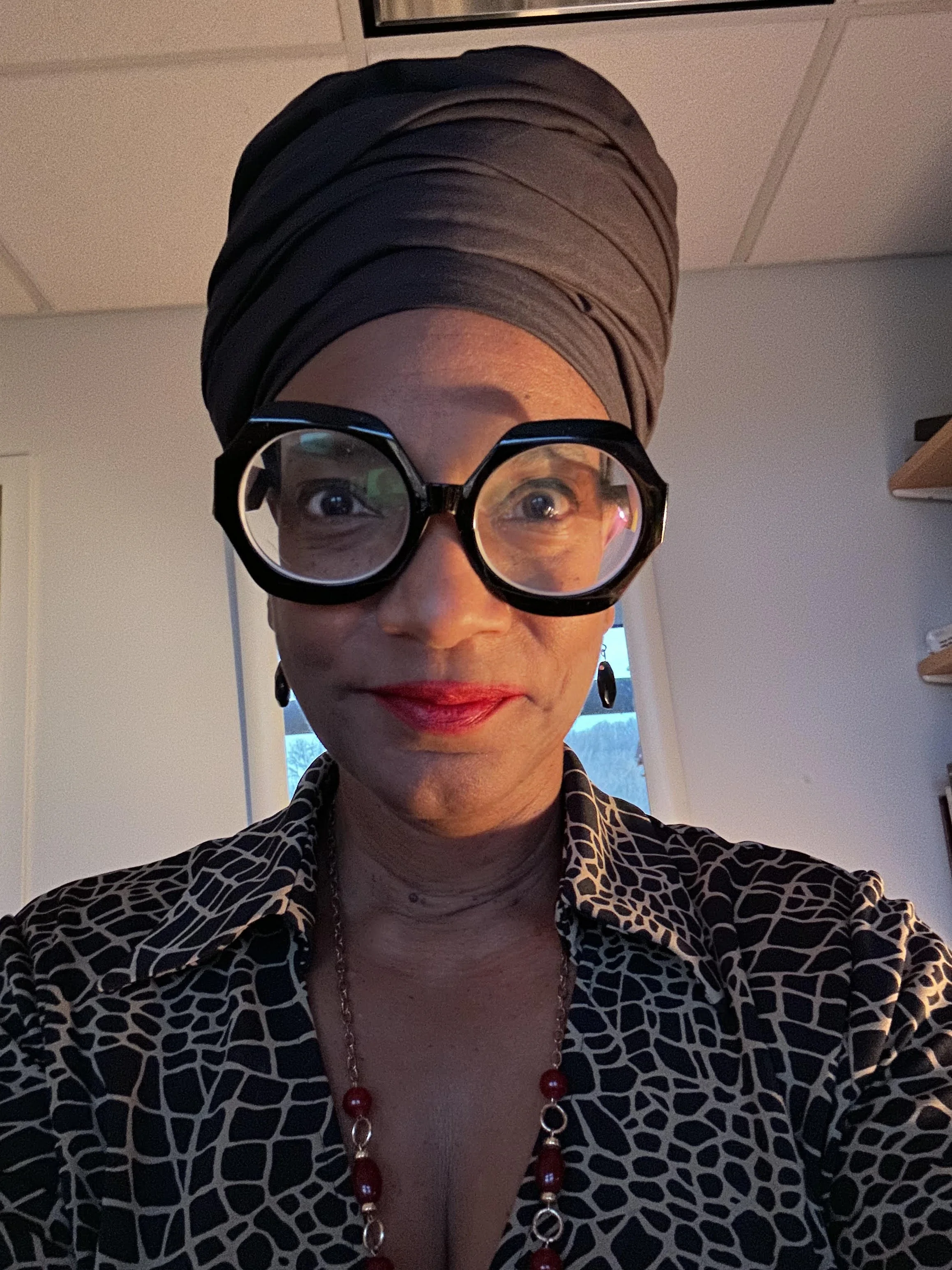 A woman wearing large, black, round glasses and a brown headwrap taking a selfie indoors. She is dressed in a patterned top with an abstract animal print and accessorized with a red bead necklace and matching earrings, with a window and part of a bookshelf in the background.
