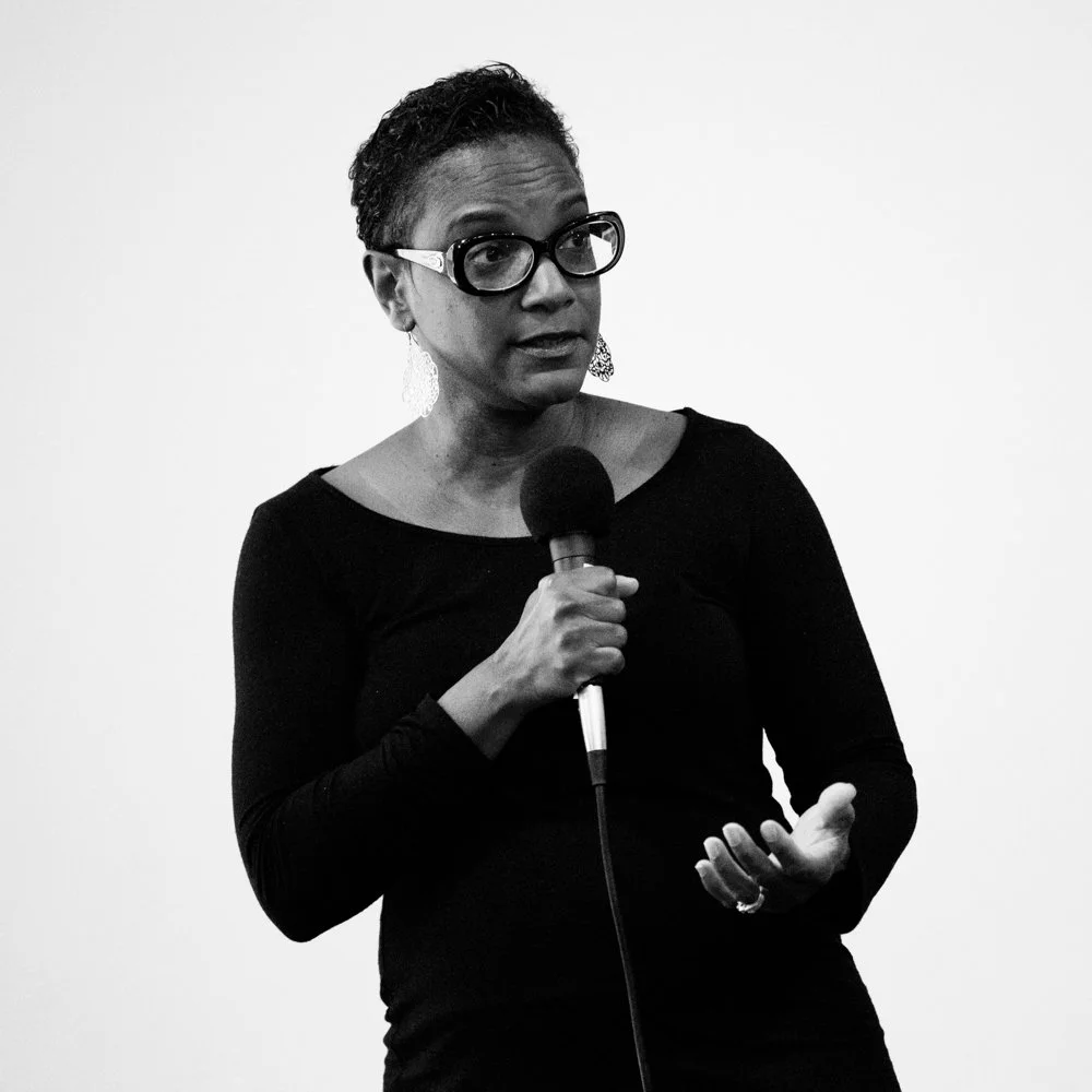 A woman with glasses and earrings holding a microphone, mid-speech, against a plain light background.