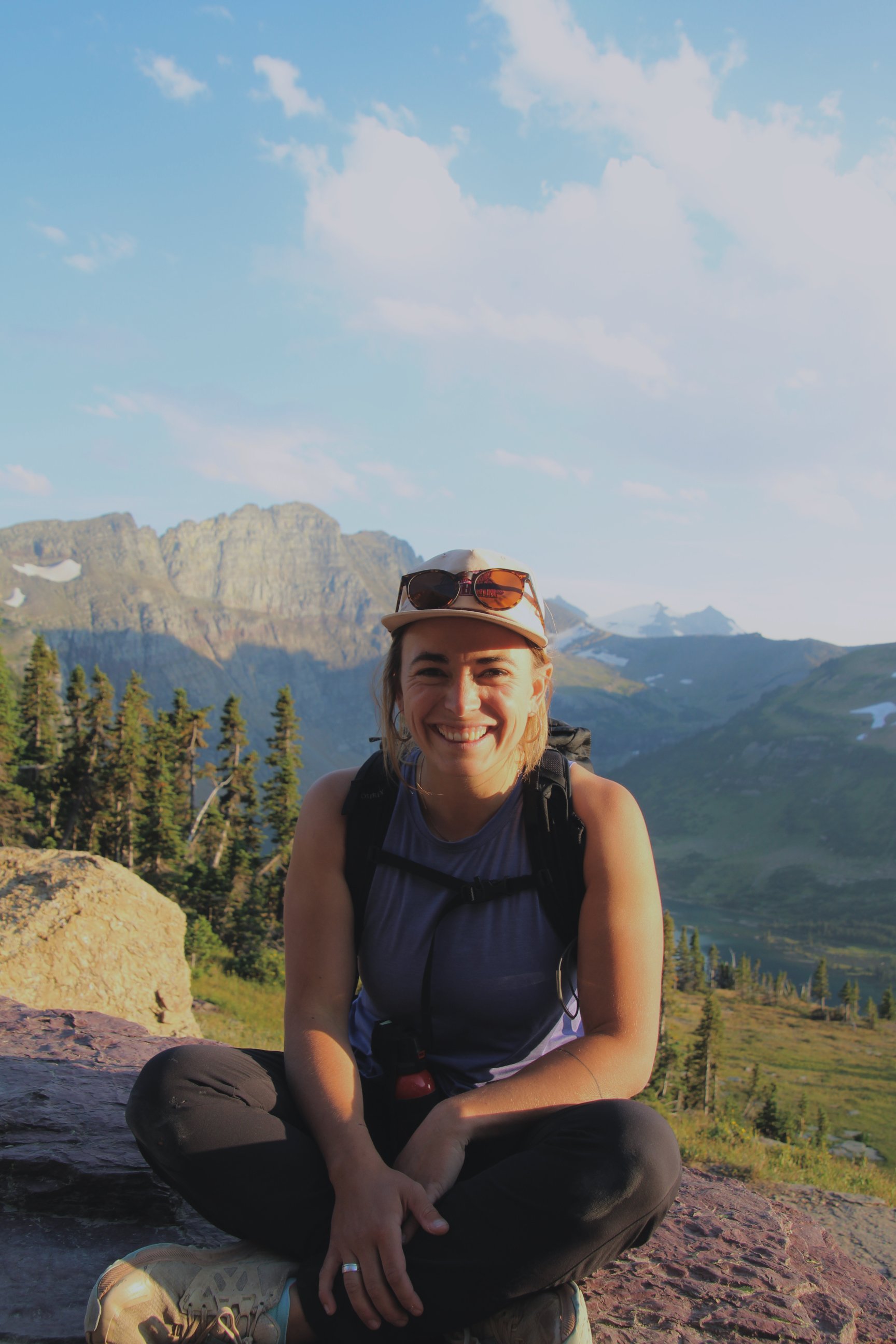 A woman smiling at the camera, sitting on a rock in a mountainous landscape with trees, mountains, and a partly cloudy sky in the background. She is wearing a hat, sunglasses, a backpack, and hiking clothes.