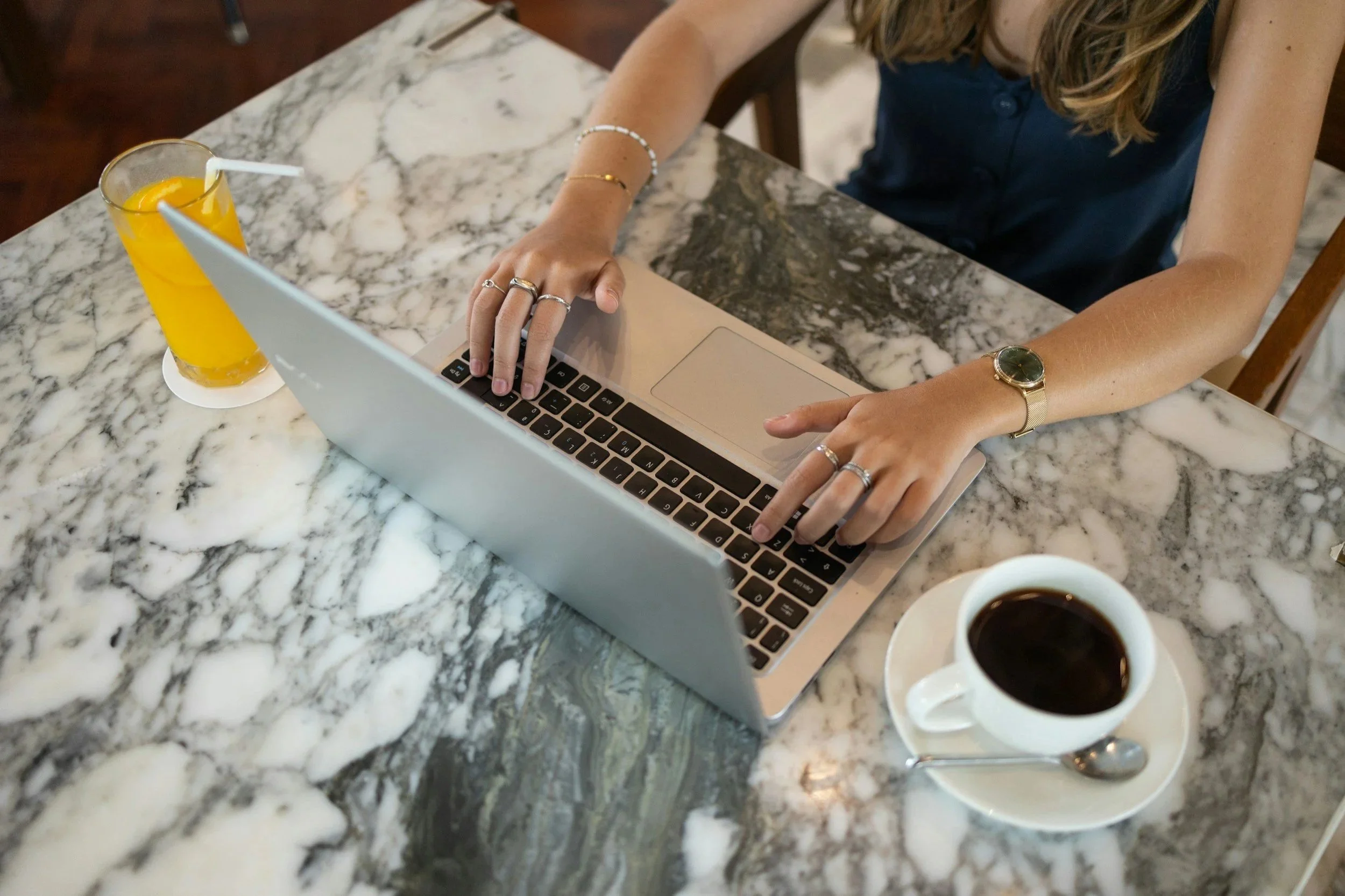 A woman sitting at a marble table working on a laptop, with a glass of orange juice and a cup of coffee in front of her.