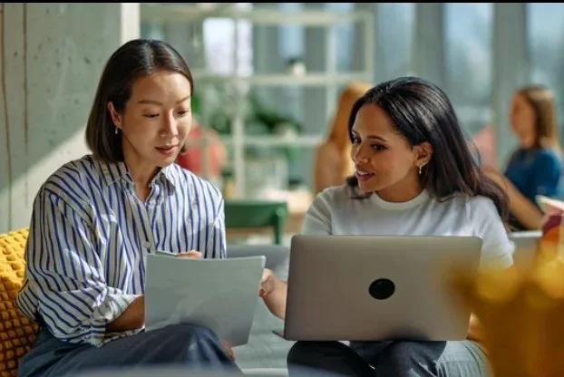 Two women sitting at a table in a modern cafe, looking at a tablet and a laptop, engaging in conversation.