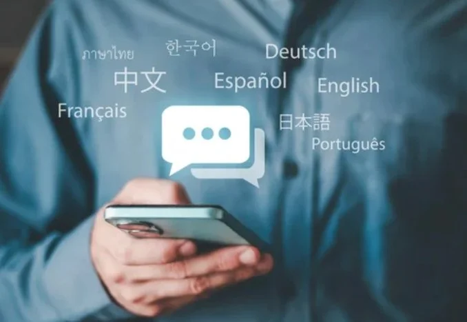Person holding a smartphone with a speech bubble icon above it surrounded by the names of various languages in different scripts, including English, Spanish, French, German, Chinese, Japanese, Korean, Portuguese, and Thai.