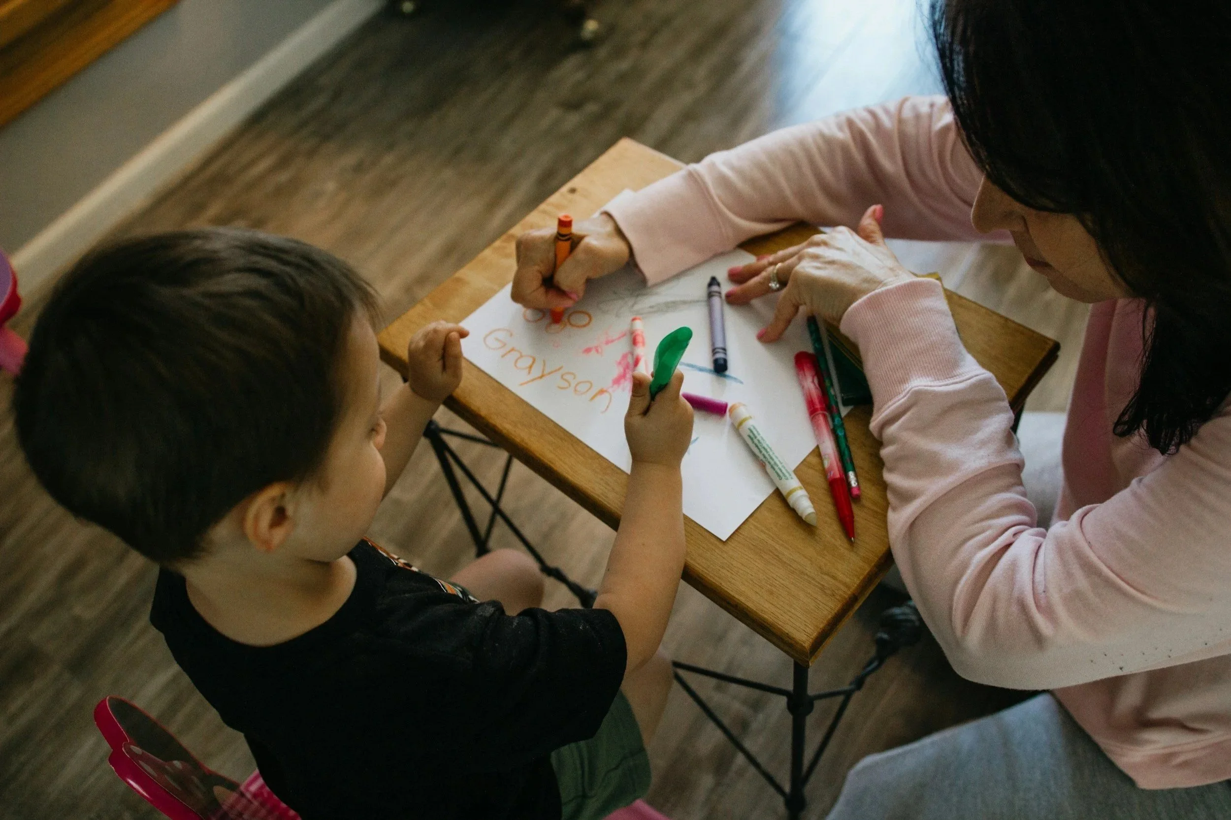 A woman helping a child with drawing or writing on a piece of paper at a wooden table, surrounded by colorful markers and crayons.