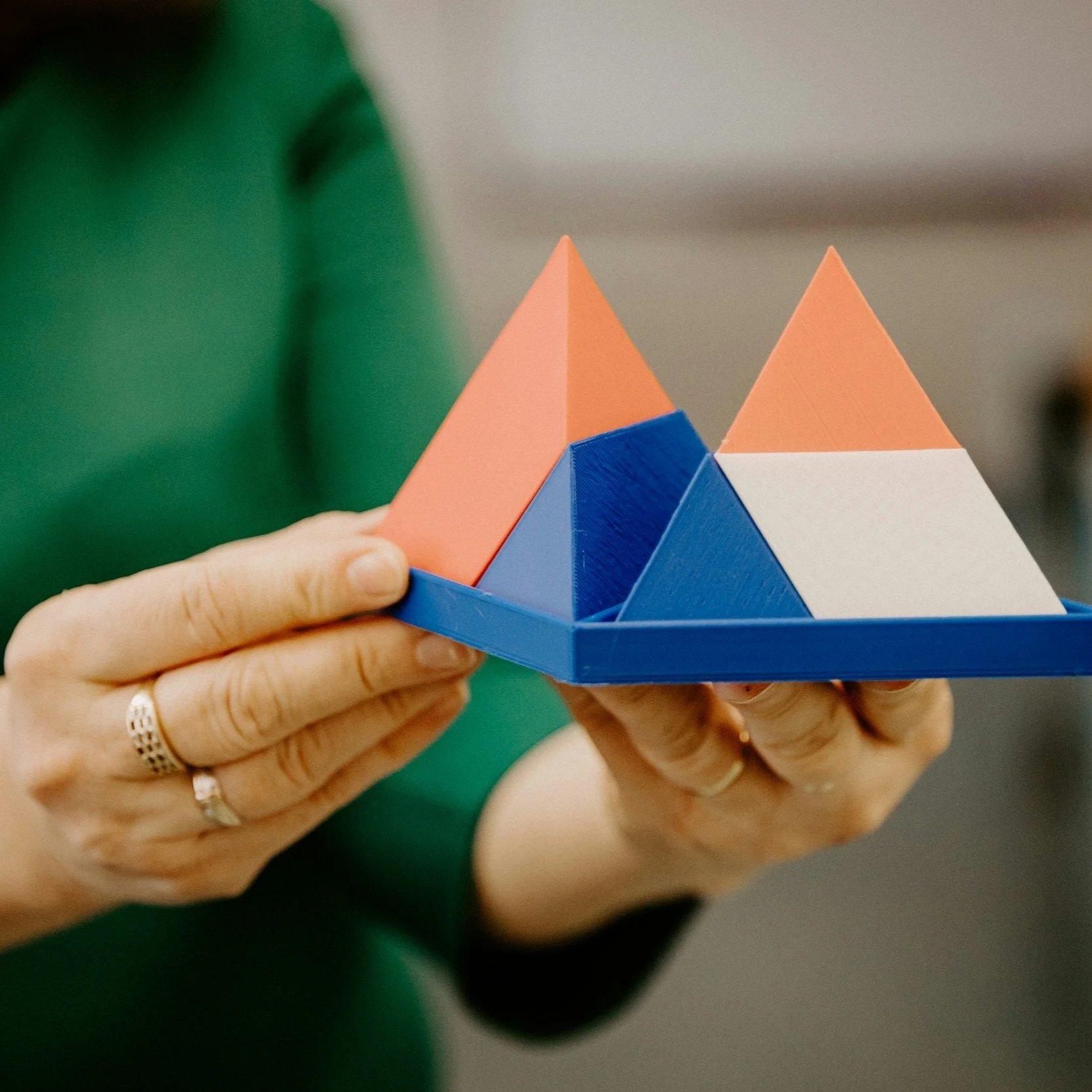 Close-up of a person holding a geometric puzzle with orange, blue, white, and pink triangular pieces, with hands wearing rings and a person in a green shirt in the background.