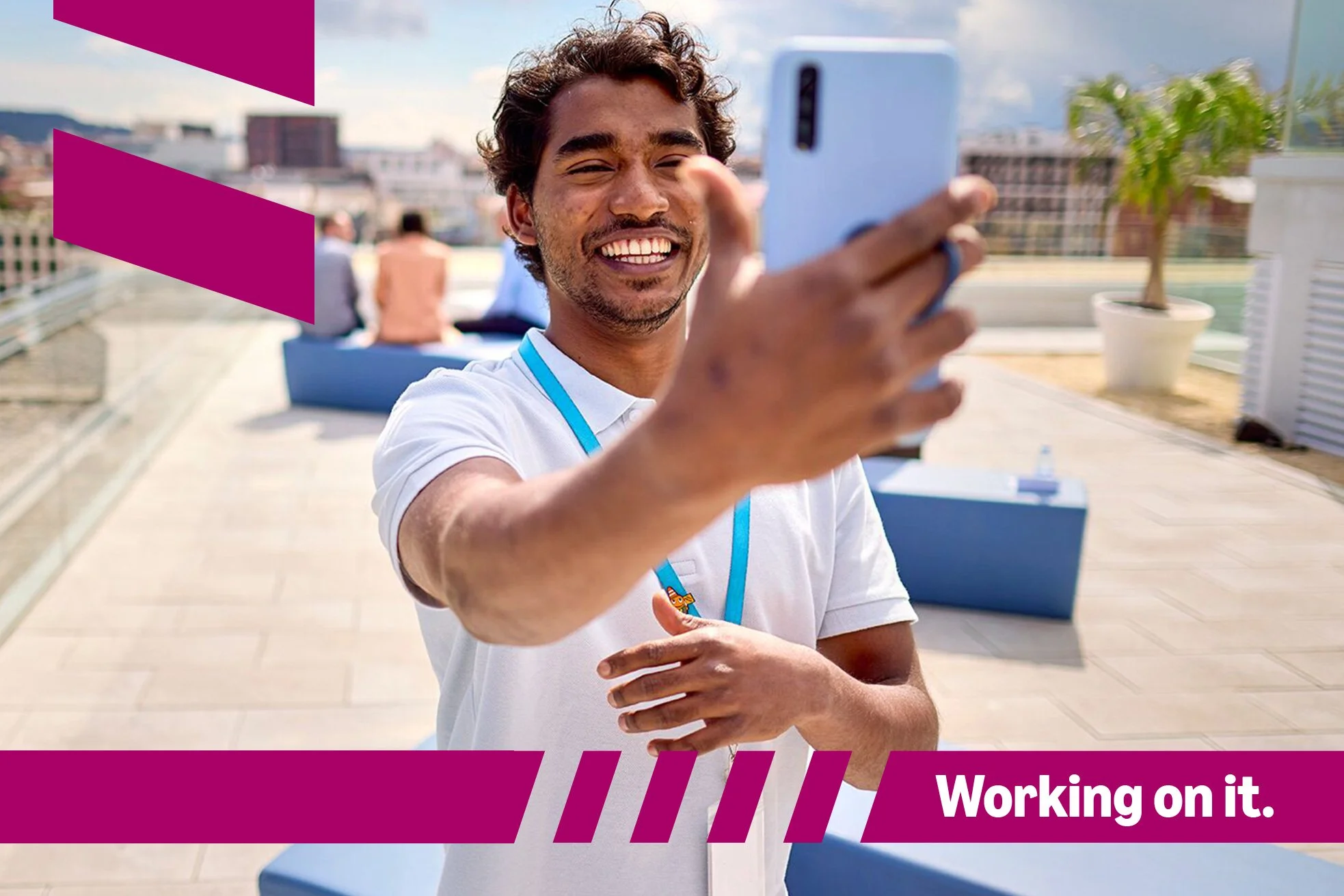 A man taking a selfie on a rooftop terrace with a city skyline in the background. He is smiling and looking at his phone, wearing a white shirt and a blue lanyard. Behind him, there are chairs, potted plants, and other people in the distance.