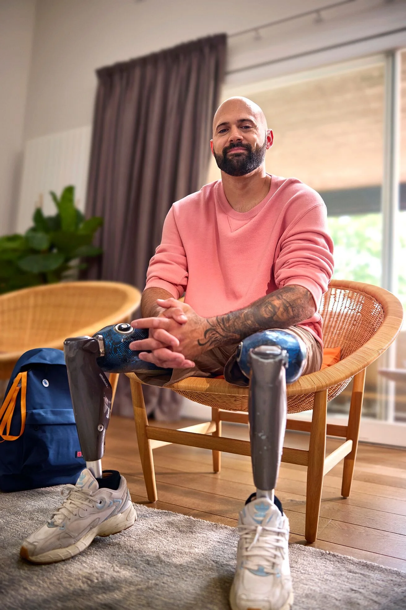 A bald man with a beard wearing a pink sweatshirt, sitting on a wooden chair with a prosthetic left arm and right leg, inside a room with large windows, brown curtains, and a green plant in the background. He is smiling with his hands clasped.