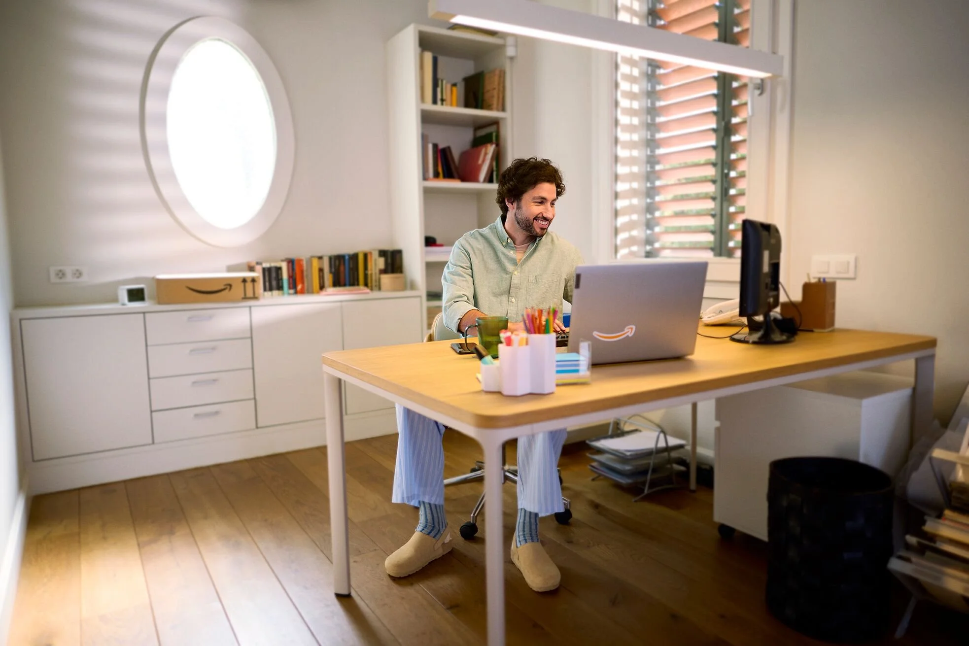 A man sitting at a desk, smiling at a laptop, in a bright, modern room with a bookshelf, window, and various office supplies.