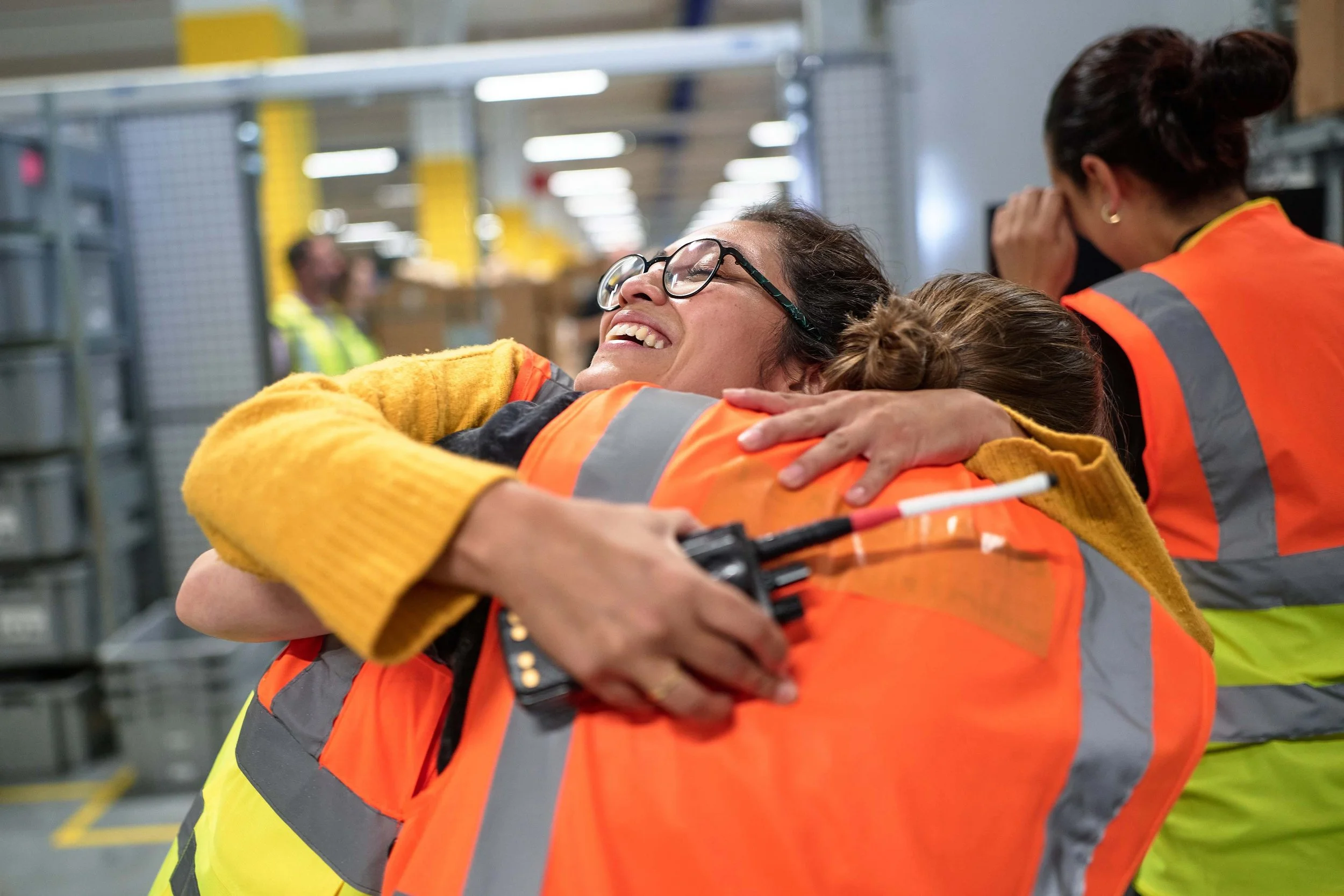 Two women in safety vests hugging each other in an industrial warehouse setting.