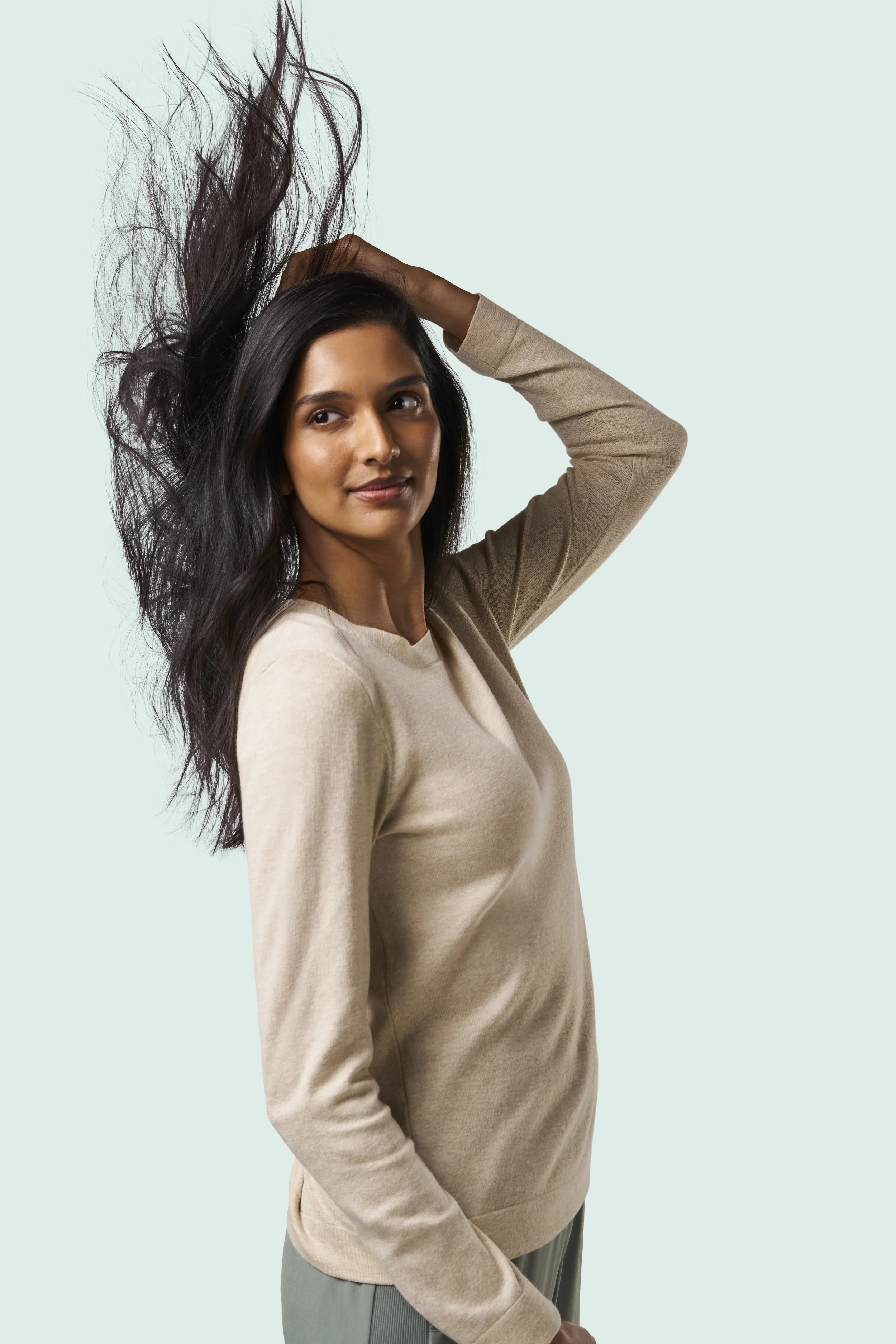 A woman with dark hair, wearing a beige sweater, has her left hand resting on her head as her hair flows up and to the left against a light mint green background.