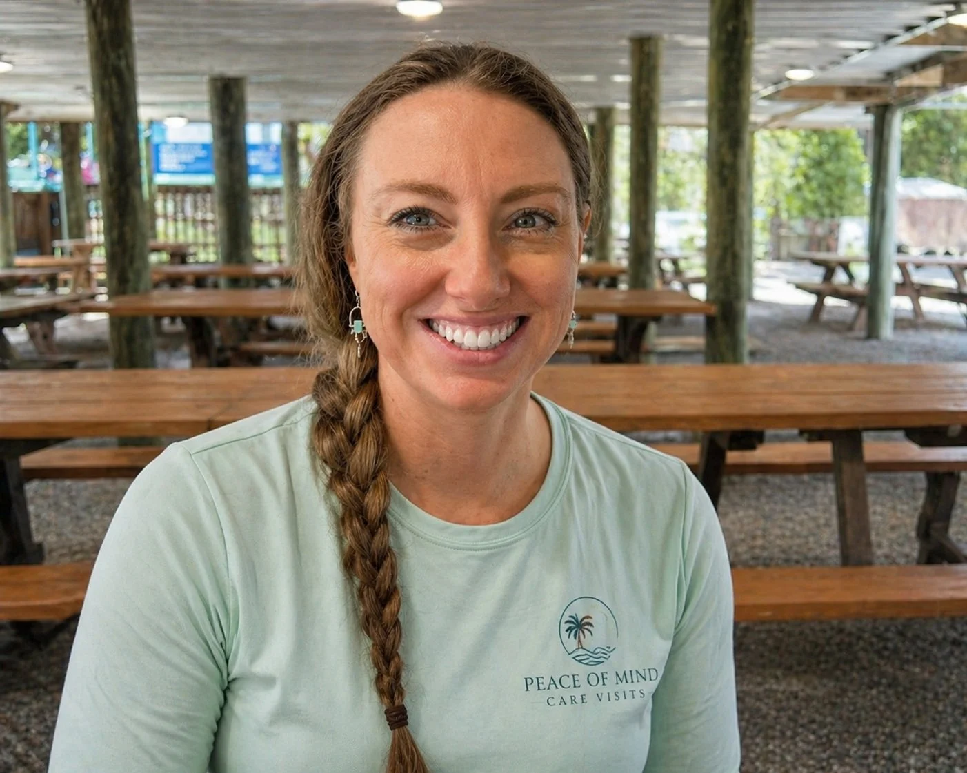 A smiling woman with a long braid, wearing earrings and a light green T-shirt with a logo, seated in an outdoor covered pavilion with wooden picnic tables.