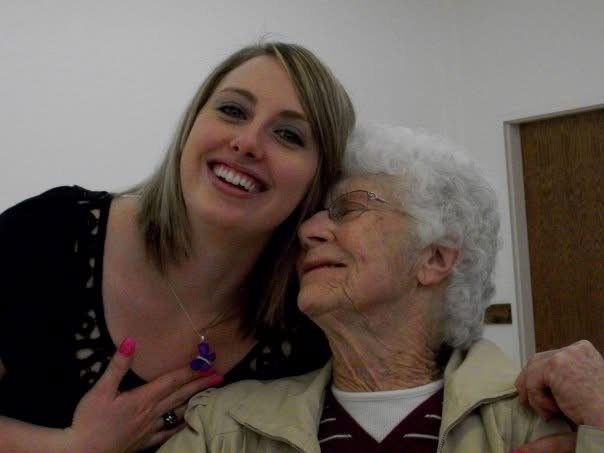 A young woman with shoulder-length blonde hair smiling and holding her hand over her chest, standing next to an elderly woman with white curly hair wearing glasses and a beige jacket, both indoors.