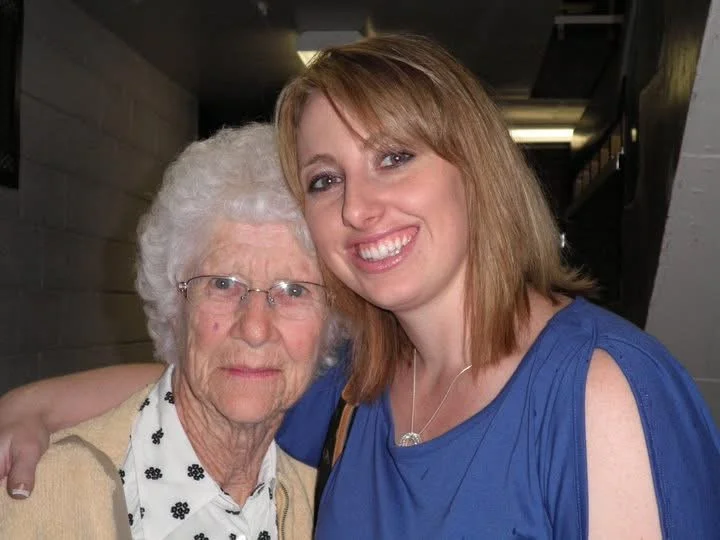 A young woman with reddish-brown hair and a blue dress taking a close-up selfie with an elderly woman with white curly hair, glasses, and a beige cardigan, inside a building.